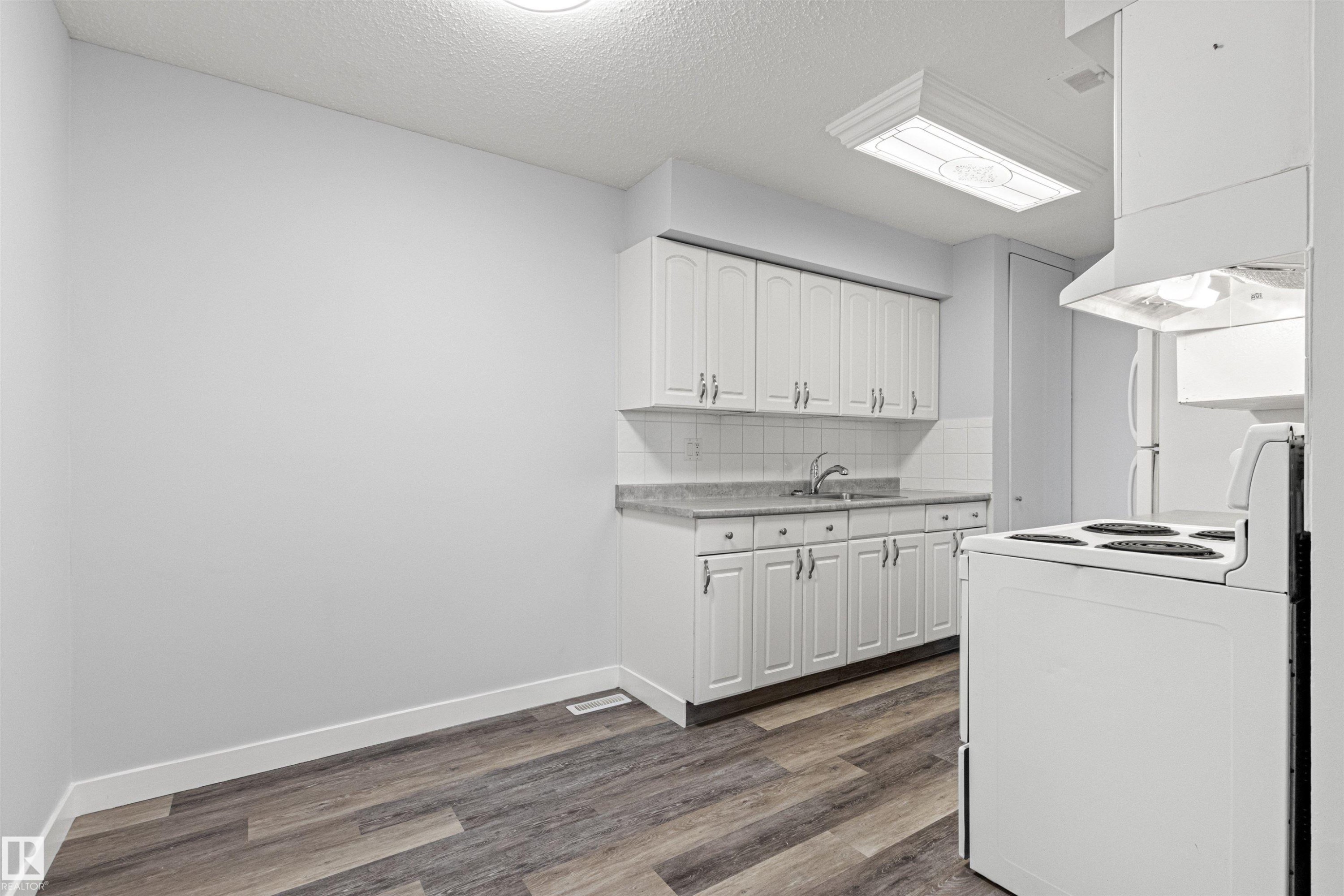 Galley kitchen with wood-finish flooring, white cabinetry, tile backsplash, and a wall-mounted range hood - #9 14230 80 St Nw, Edmonton, AB - Indoor Photo Showing Kitchen