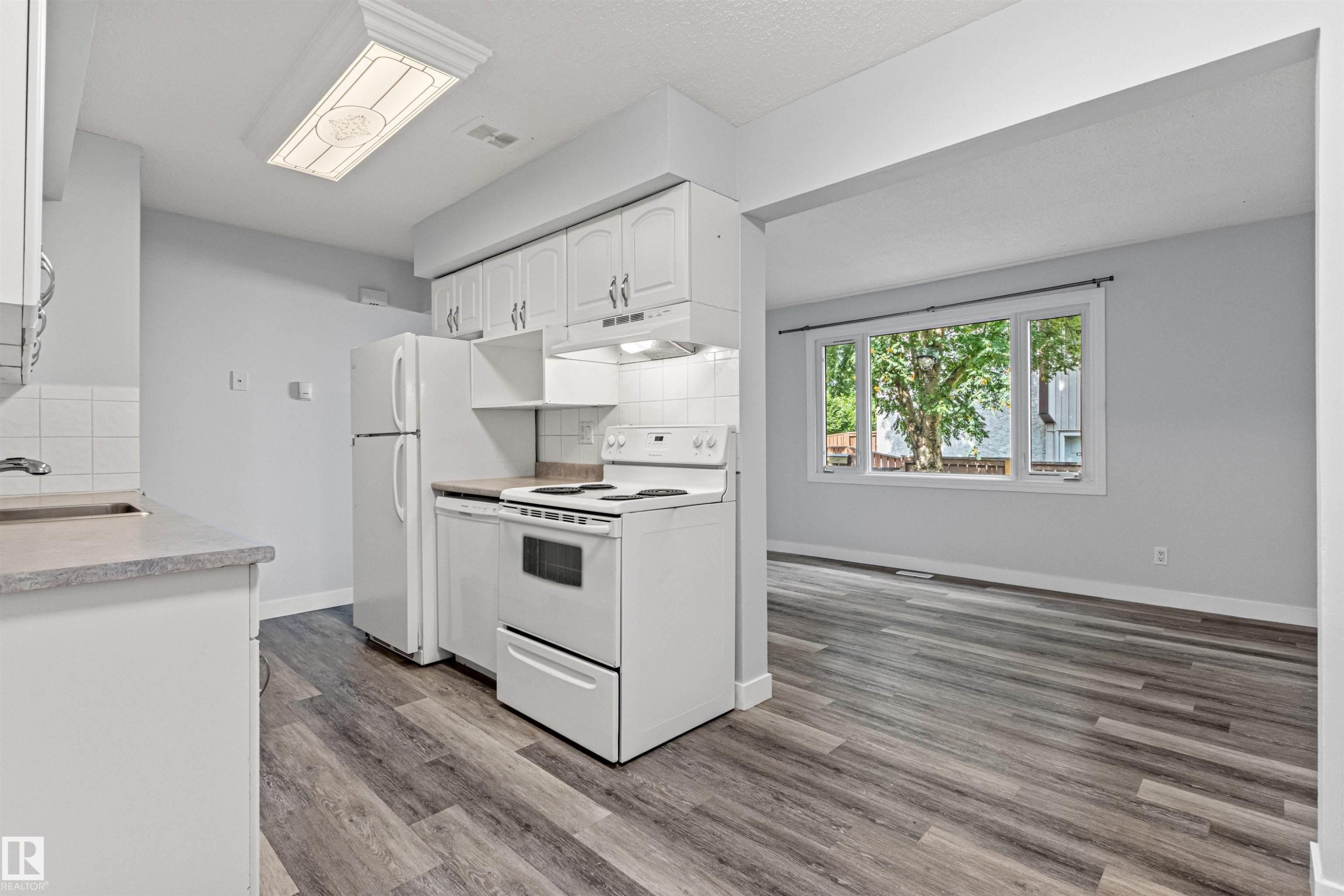 Open-concept kitchen and living area featuring white cabinetry, stainless steel appliances, wood-finish flooring, and abundant natural light from multi-pane windows overlooking mature landscaping - #9 14230 80 St Nw, Edmonton, AB - Indoor Photo Showing Kitchen