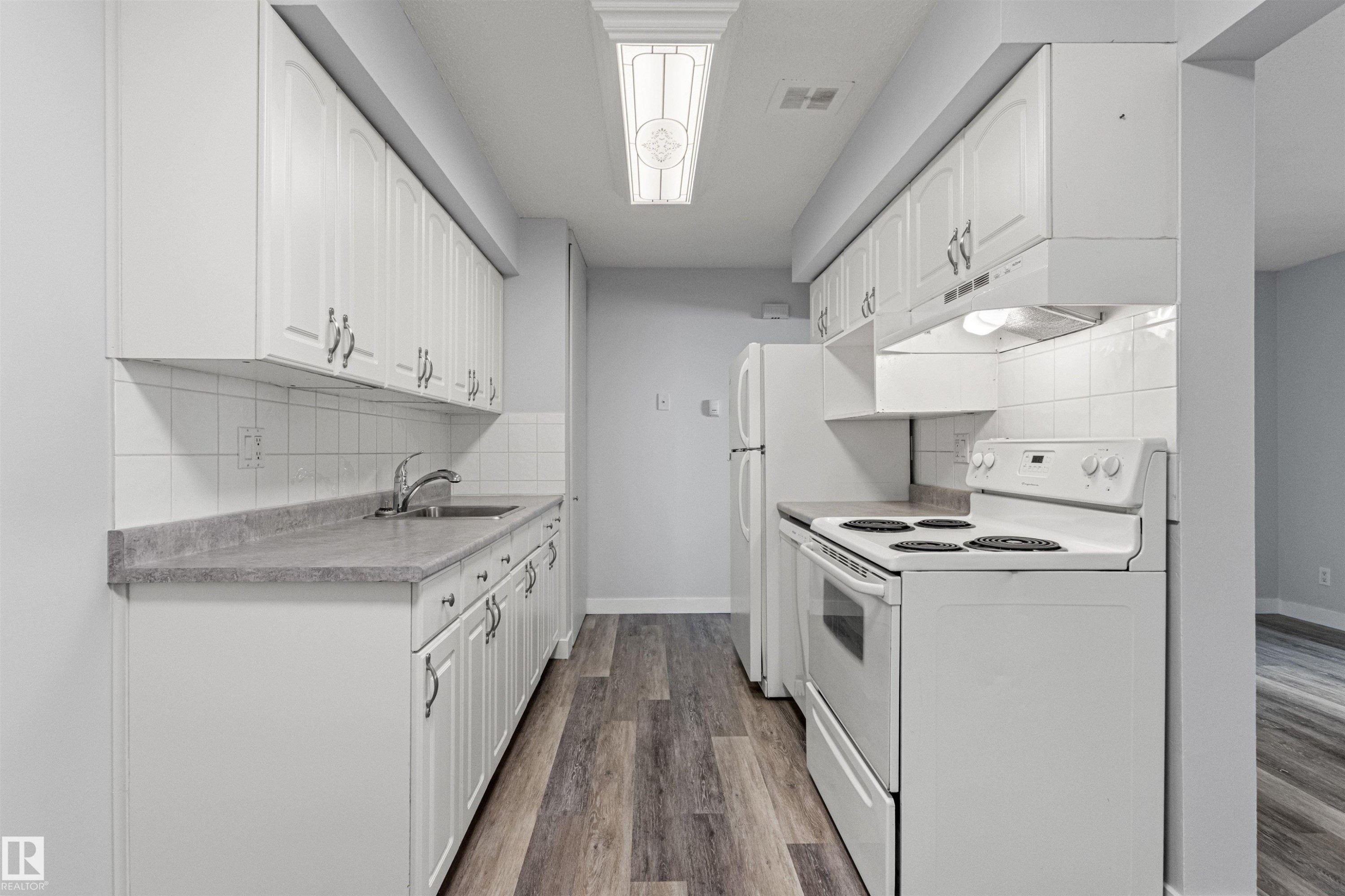 Galley-style kitchen featuring white cabinetry with chrome hardware, laminate countertops, subway tile backsplash, electric range with cooktop, refrigerator, and wood-finish flooring throughout - #9 14230 80 St Nw, Edmonton, AB - Indoor Photo Showing Kitchen