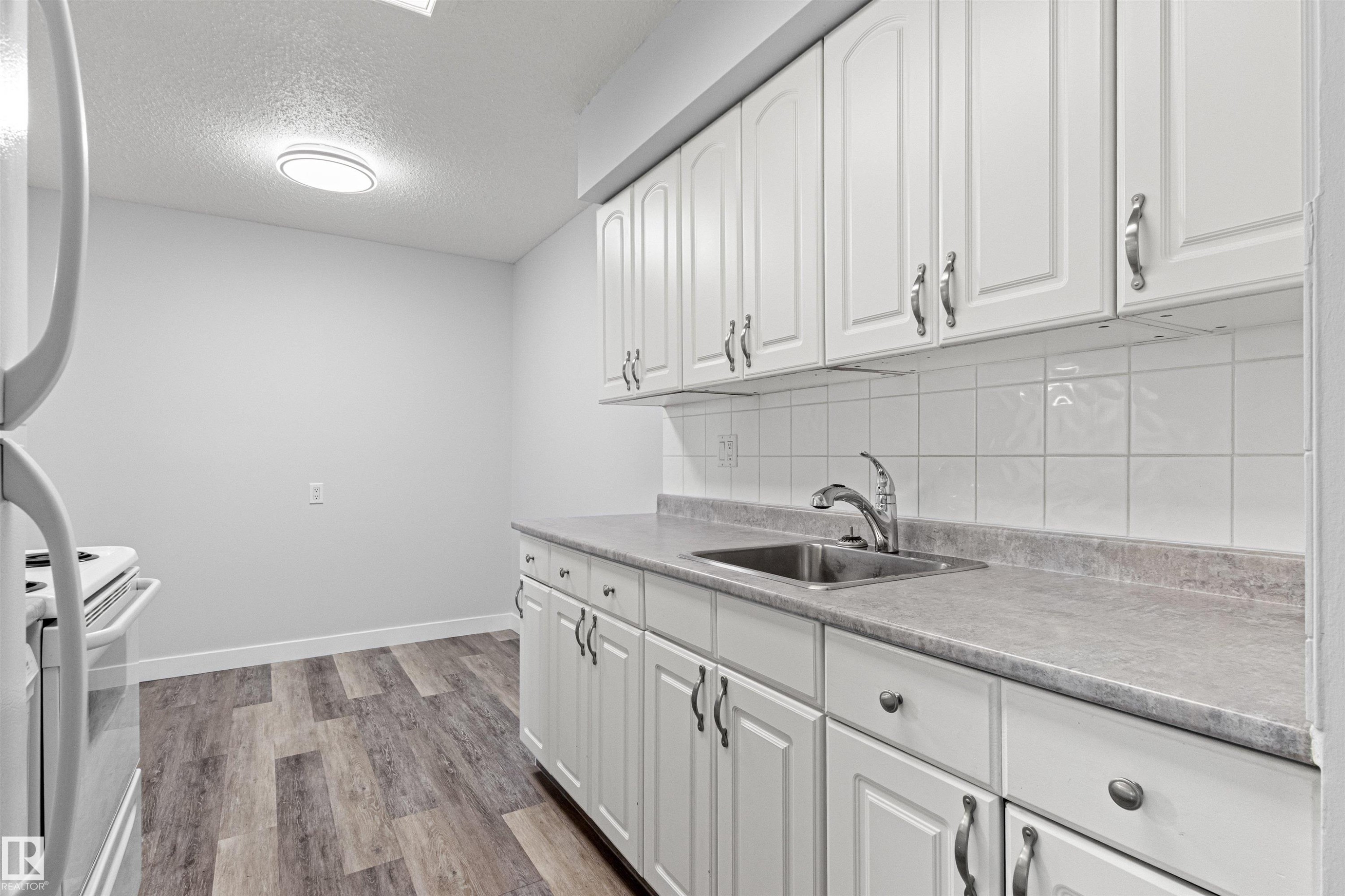 Wood-finish flooring with white baseboards, white shaker-style cabinetry, square tile backsplash, and a stainless steel sink with a gooseneck faucet - #9 14230 80 St Nw, Edmonton, AB - Indoor Photo Showing Kitchen