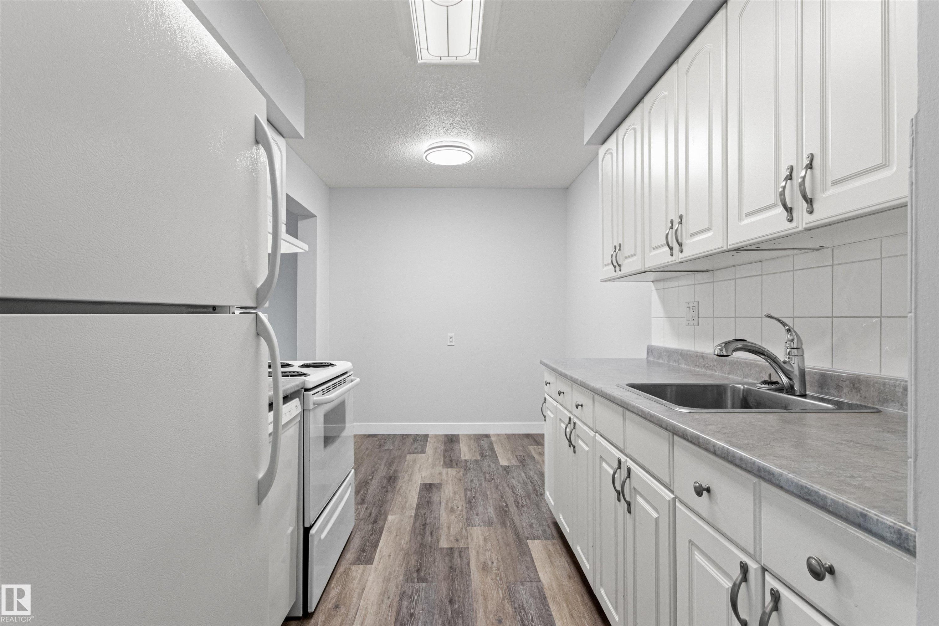 Galley kitchen featuring white cabinetry with hardware accents, stainless steel appliances including a gas range and refrigerator, gray countertops, subway tile backsplash, and wood-finish flooring throughout - #9 14230 80 St Nw, Edmonton, AB - Indoor Photo Showing Kitchen