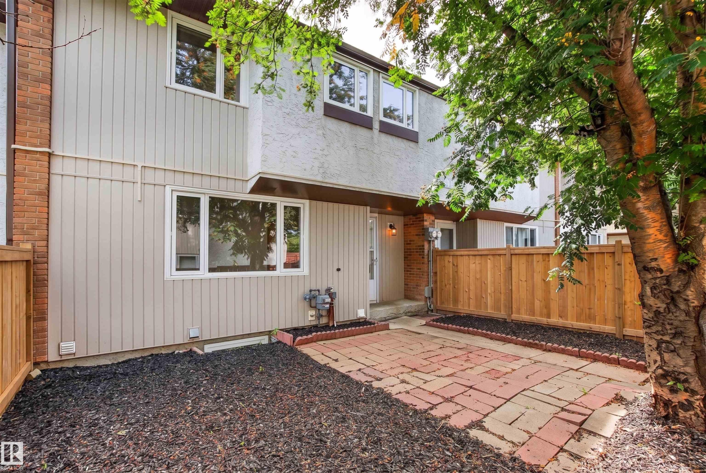Modern townhome exterior featuring a private patio with brick and gravel hardscaping, wooden privacy fencing, contemporary siding with metal accents, large multi-pane windows, and mature landscaping with established trees providing natural screening - #9 14230 80 St Nw, Edmonton, AB - Outdoor With Exterior