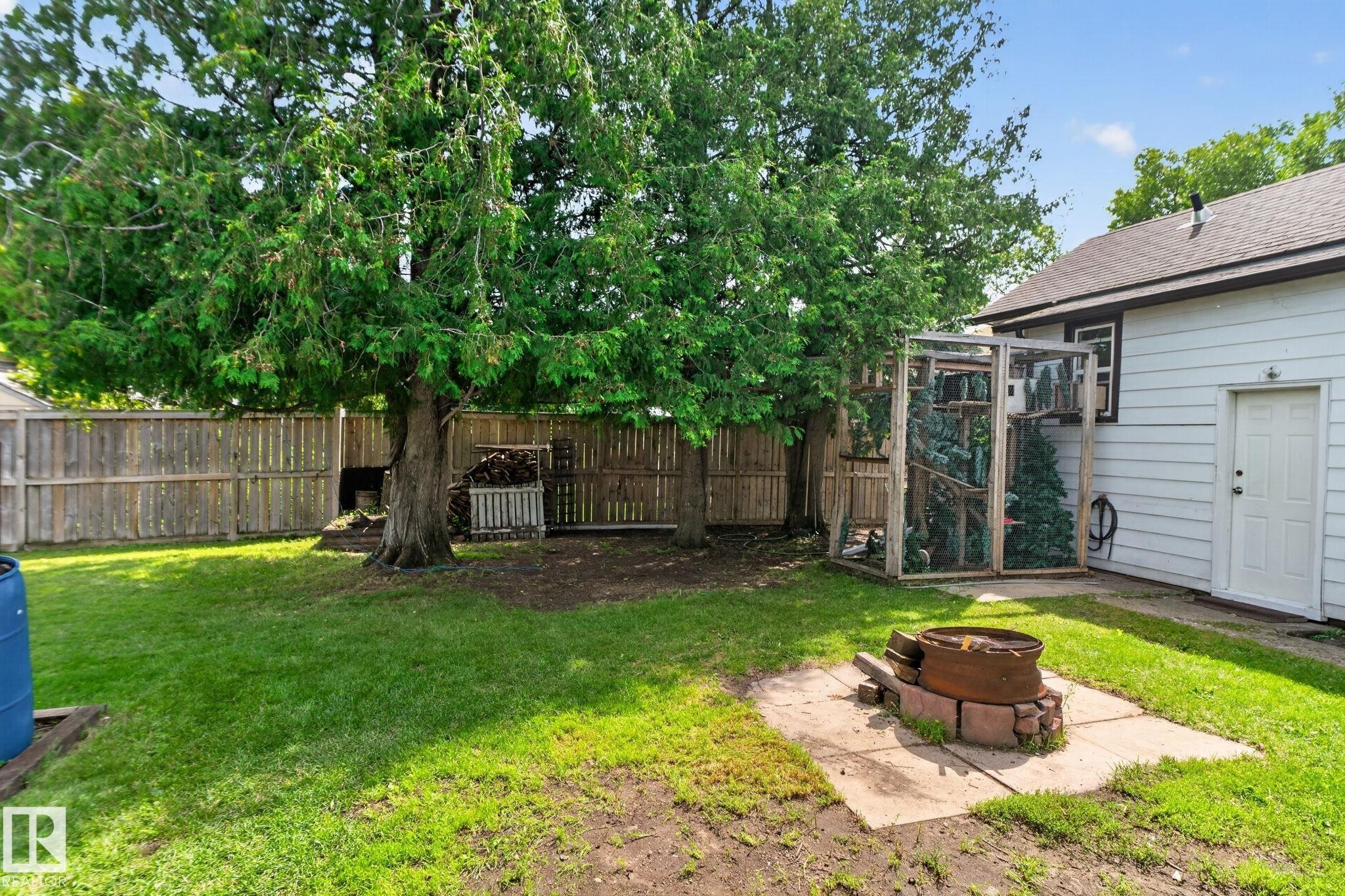 Expansive backyard featuring a manicured lawn, established trees, wood fencing, and a built-in fire pit on a concrete pad - 9921 109 Street, Fort Saskatchewan, AB - Outdoor