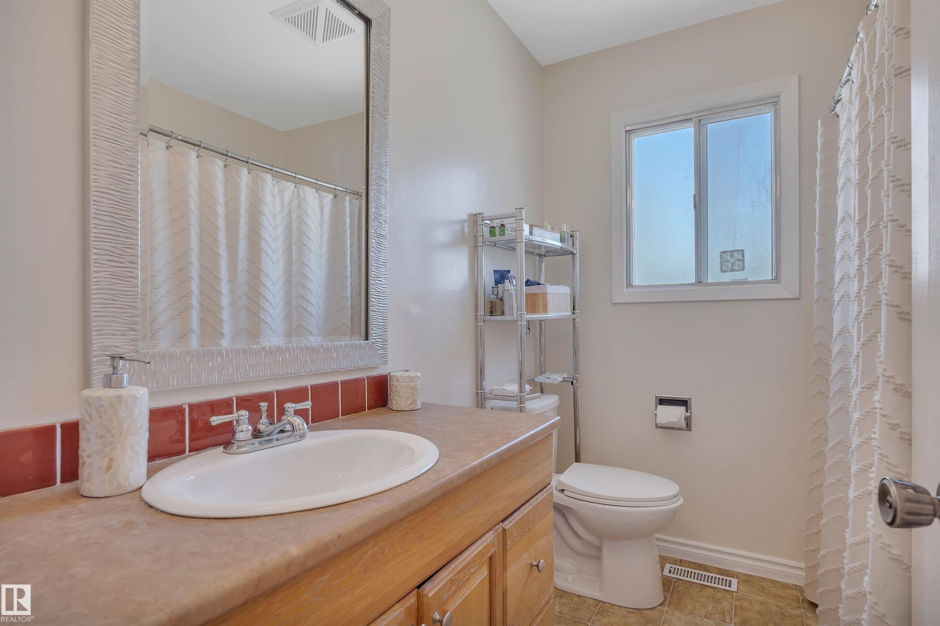 Bathroom featuring a wood-finish vanity with an oval sink, a large framed mirror, terracotta-toned tile backsplash, and a window providing natural light - 5411 44 Ave, Wetaskiwin, AB - Indoor Photo Showing Bathroom