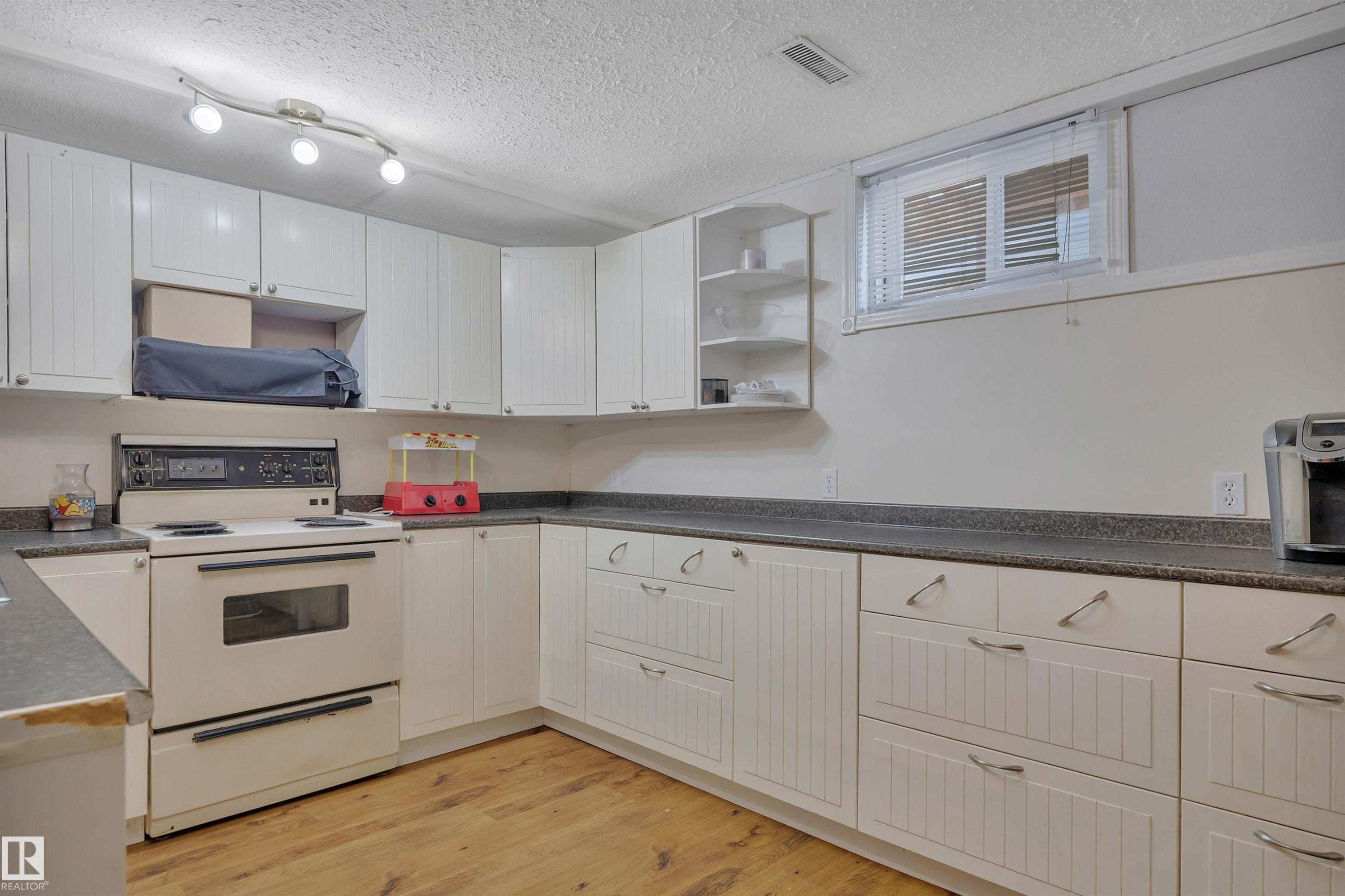 Kitchen featuring extensive white cabinetry, wood-finish flooring, a built-in range, dark countertops, and a window with blinds - 5411 44 Ave, Wetaskiwin, AB - Indoor Photo Showing Kitchen