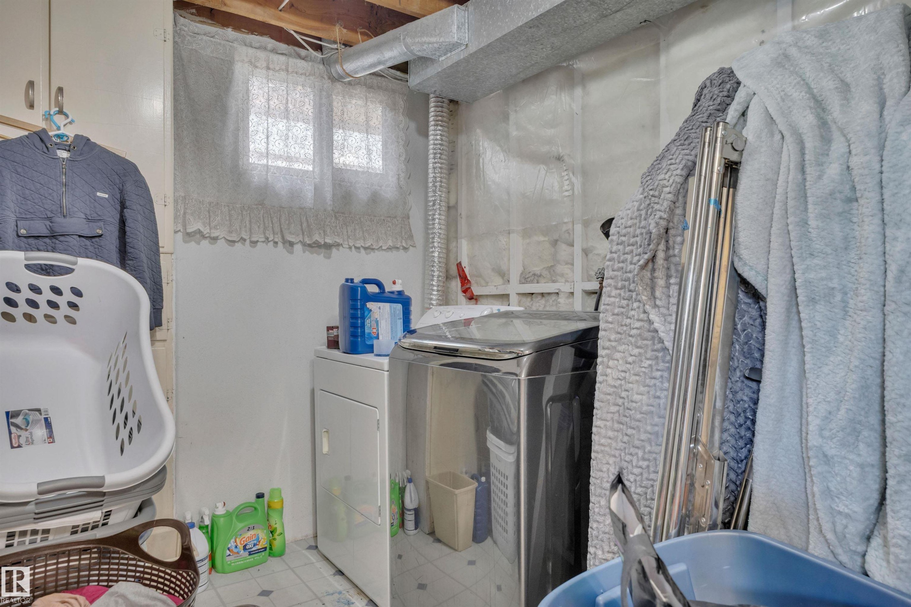 Laundry area with a window featuring lace curtains, exposed ceiling joists, and a visible dryer vent - 5411 44 Ave, Wetaskiwin, AB - Indoor