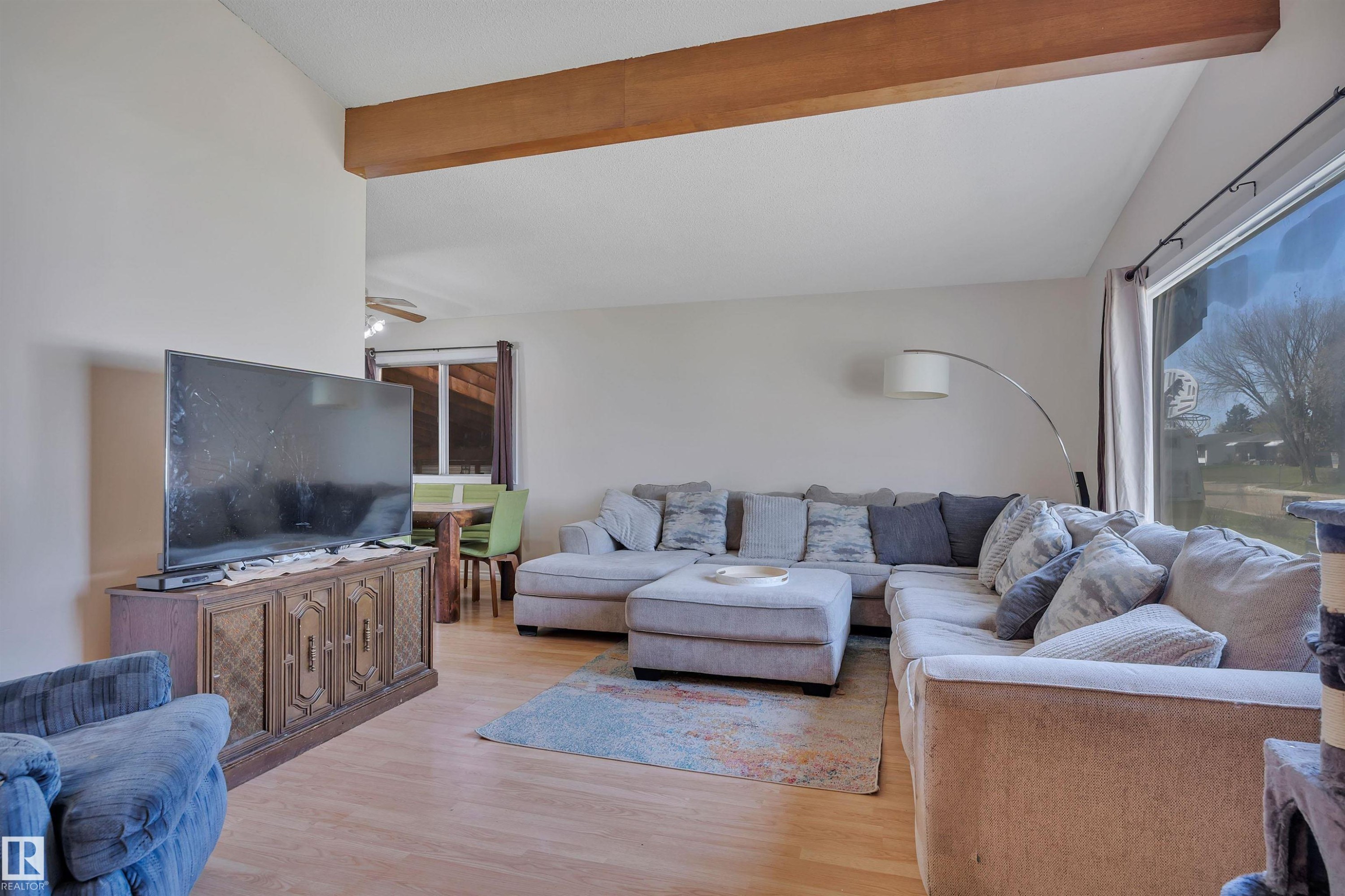 Living area featuring light wood-finish flooring, a prominent wooden ceiling beam, and a large window with vertical blinds - 5411 44 Ave, Wetaskiwin, AB - Indoor Photo Showing Living Room