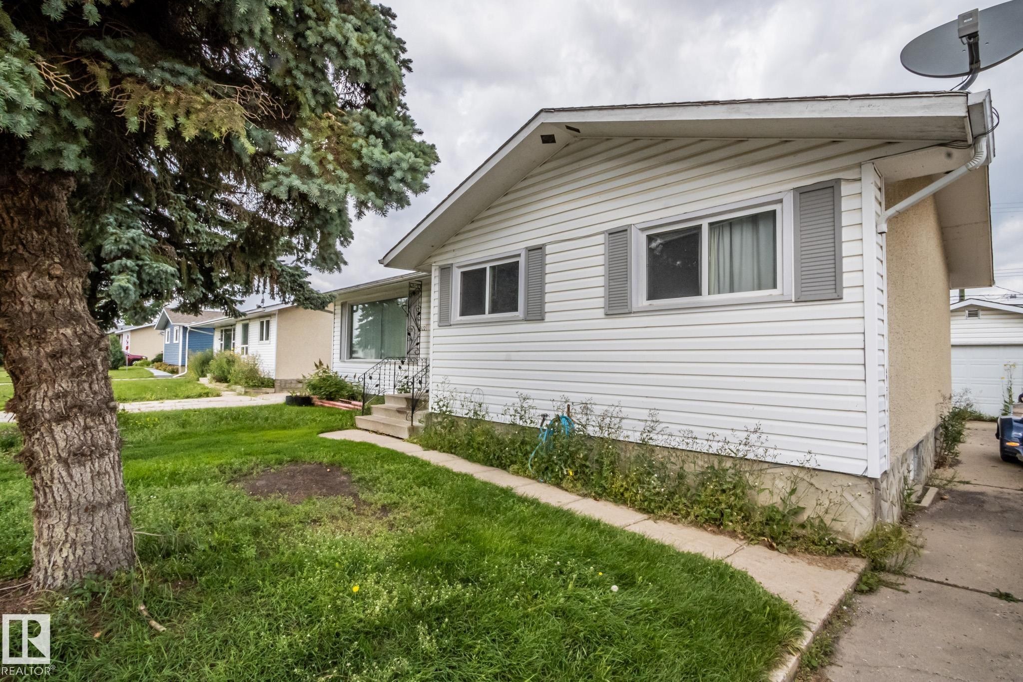 White siding exterior, gray louvered shutters, concrete walkway, front porch with wrought iron railing, mature tree - 4322 49 Ave, St. Paul Town, AB - Outdoor