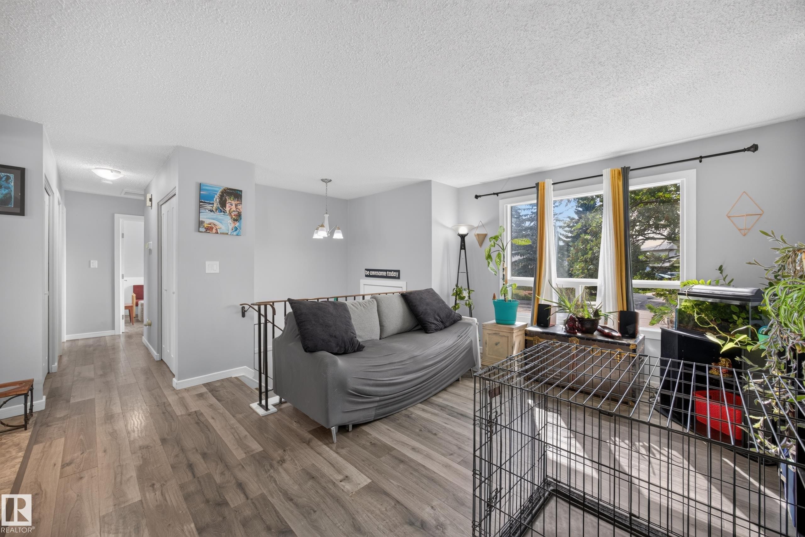 Bright living area showcasing wood-finish flooring, large multi-pane windows with natural light, gray upholstered seating, pendant lighting, and access to outdoor space with mature landscaping visible beyond - 156 Clareview Road Nw, Edmonton, AB - Indoor Photo Showing Living Room