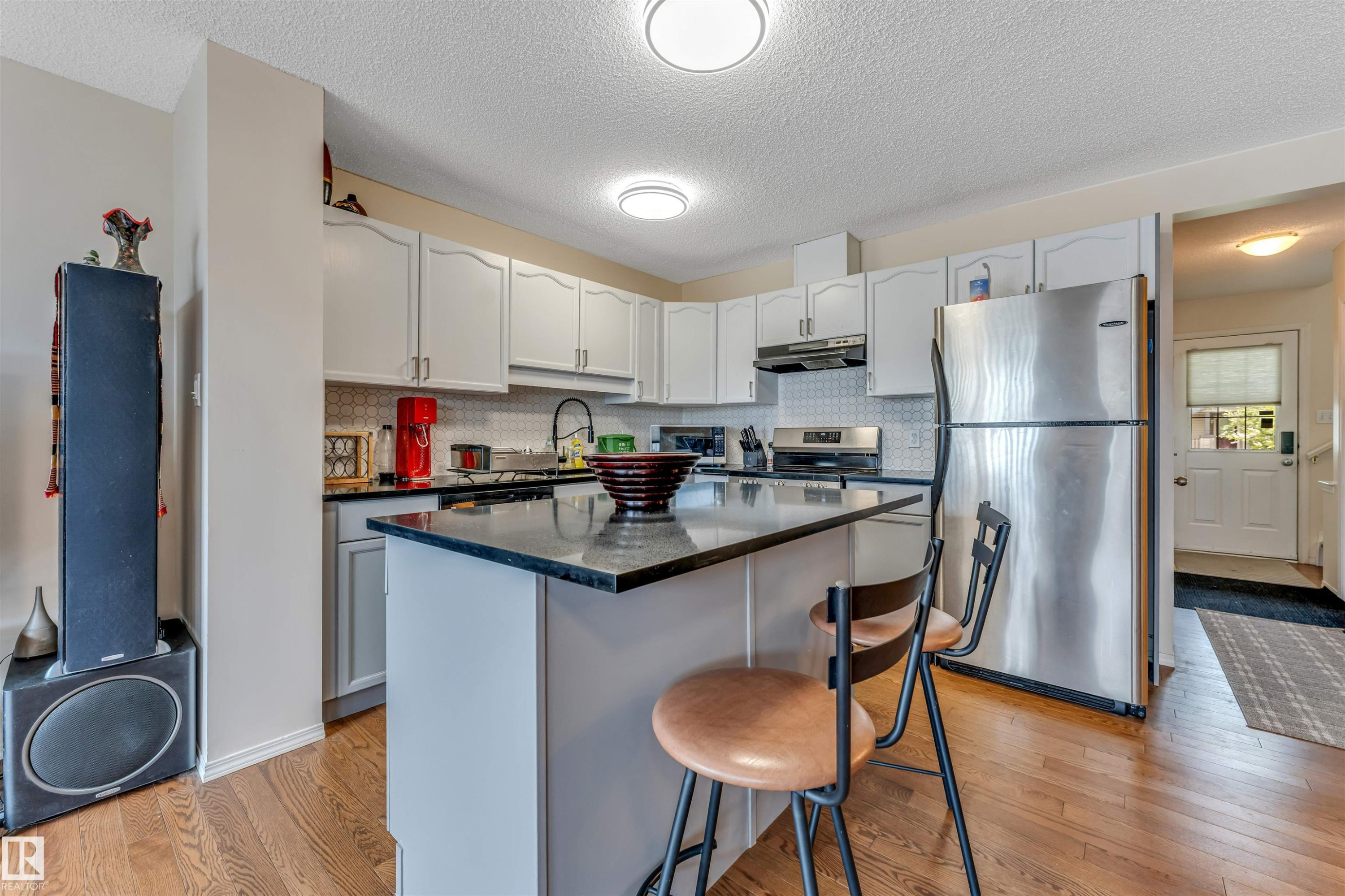 Kitchen featuring an island with a dark countertop, white cabinetry, stainless steel appliances, a white tile backsplash, and wood-finish flooring - 3032 31 Avenue Nw, Edmonton, AB - Indoor Photo Showing Kitchen With Double Sink