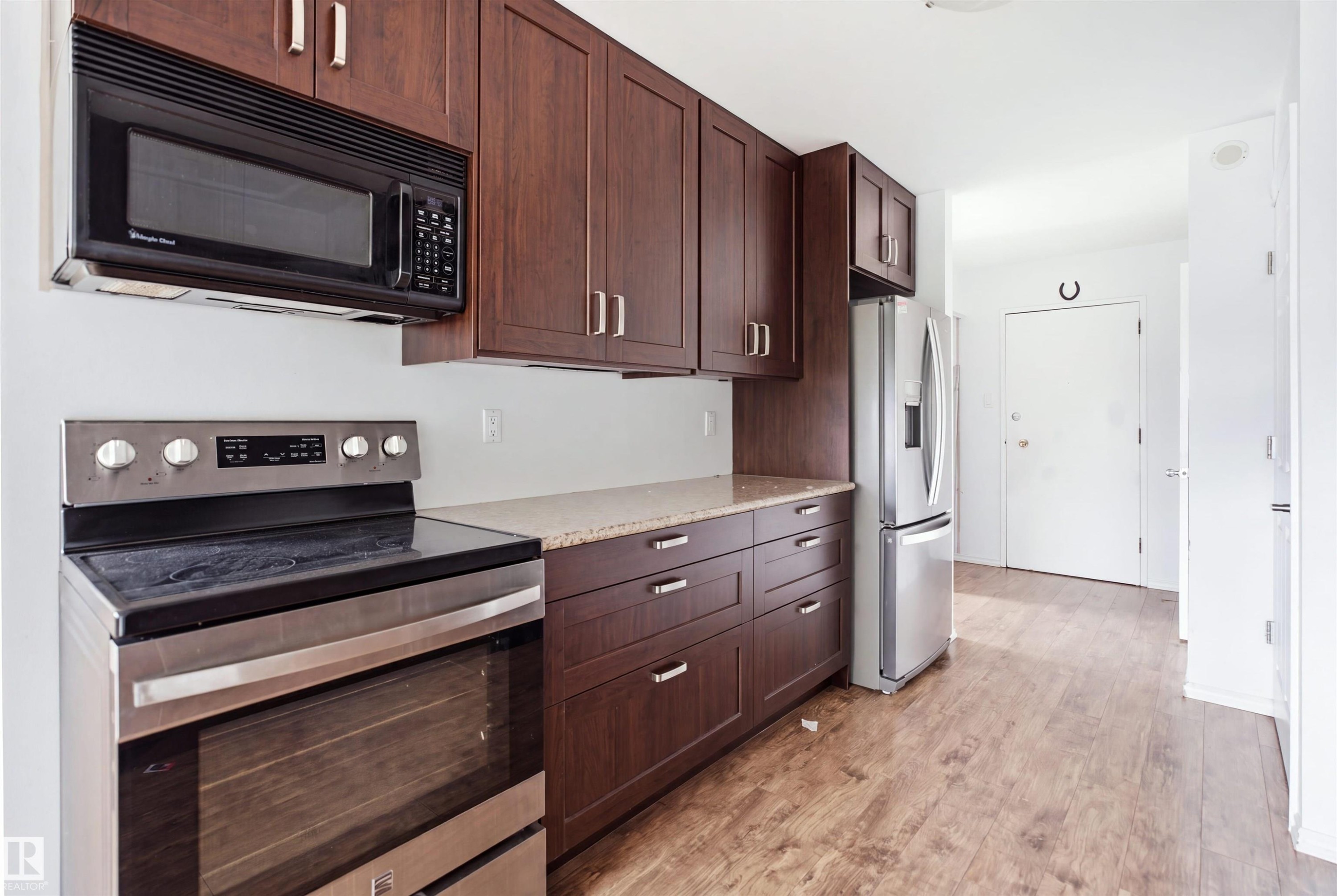 Modern kitchen featuring rich wood cabinetry with stainless steel hardware, black smooth-top electric range with matching microwave hood, white refrigerator, and dark countertops - 7825 159 Street Nw, Edmonton, AB - Indoor Photo Showing Kitchen