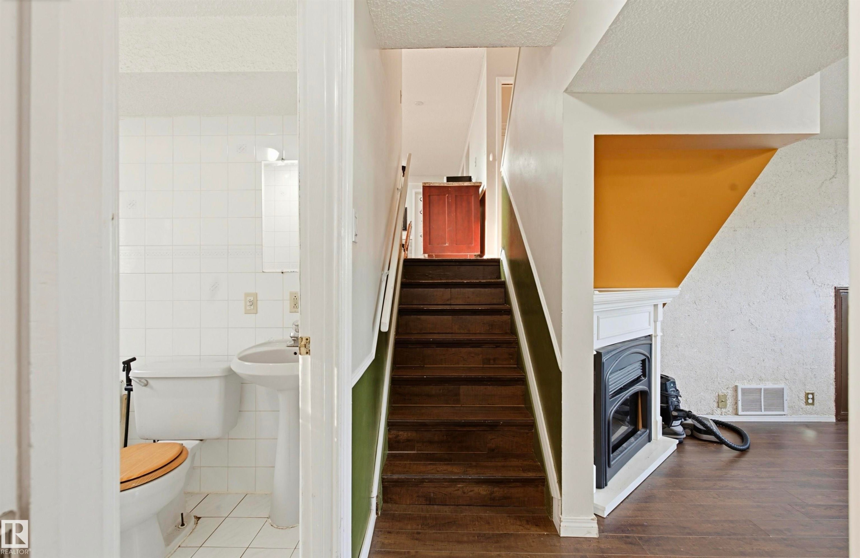 Wood-finish flooring, white tiled bathroom, pedestal sink, white toilet, staircase with wood treads, and white fireplace surround with a dark insert - 13604 90A Street Nw, Edmonton, AB - Indoor Photo Showing Other Room