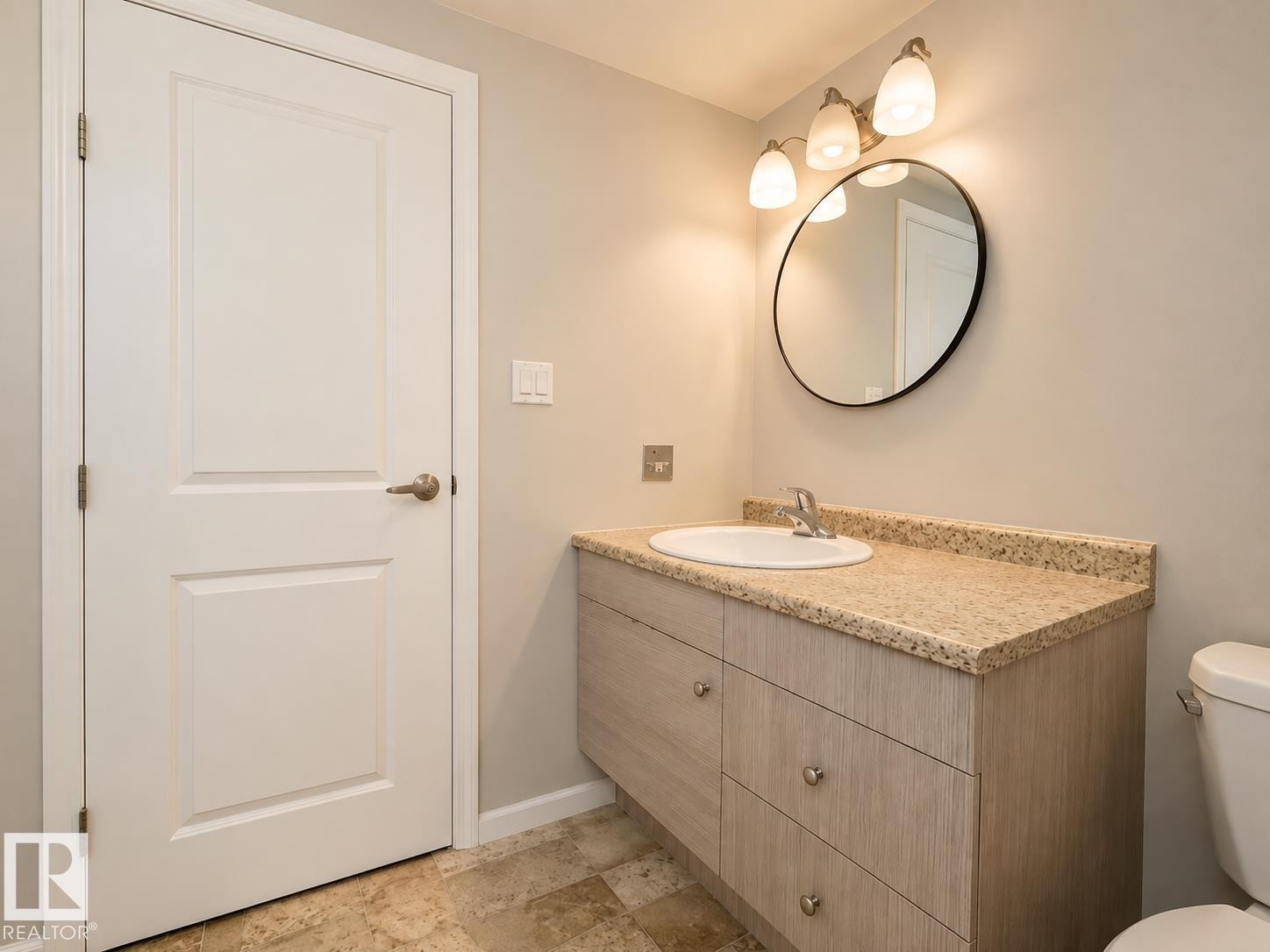 White panel door, light wood-finish vanity with drawer storage, integrated sink, stone-finish countertop, and circular mirror with three-light fixture - #31 11255 31 Av Nw, Edmonton, AB - Indoor Photo Showing Bathroom