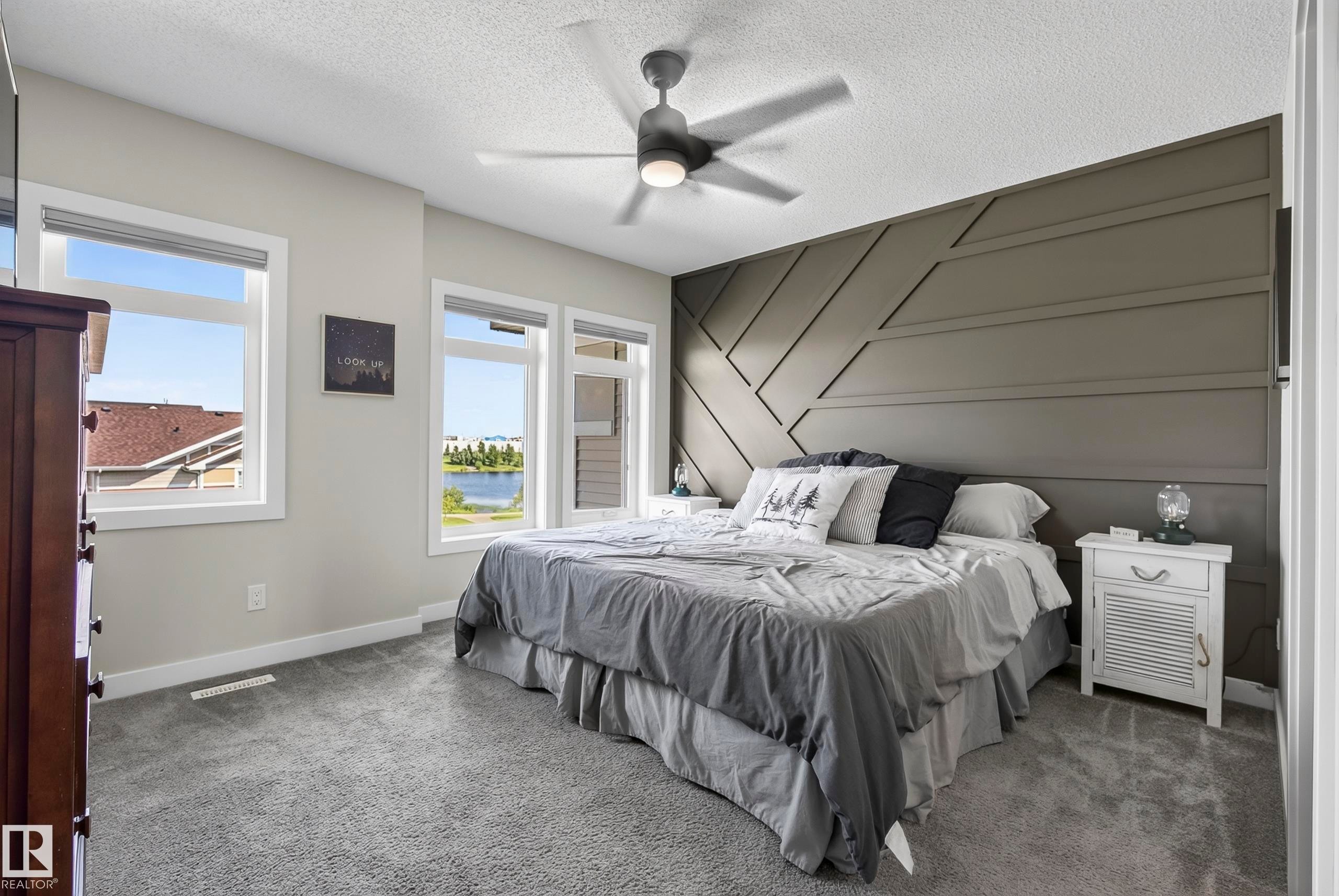 Accent wall with geometric paneling, multiple windows with a water backdrop, ceiling fan with integrated lighting, and carpeted flooring - 45 Joyal Way, St. Albert, AB - Indoor Photo Showing Bedroom