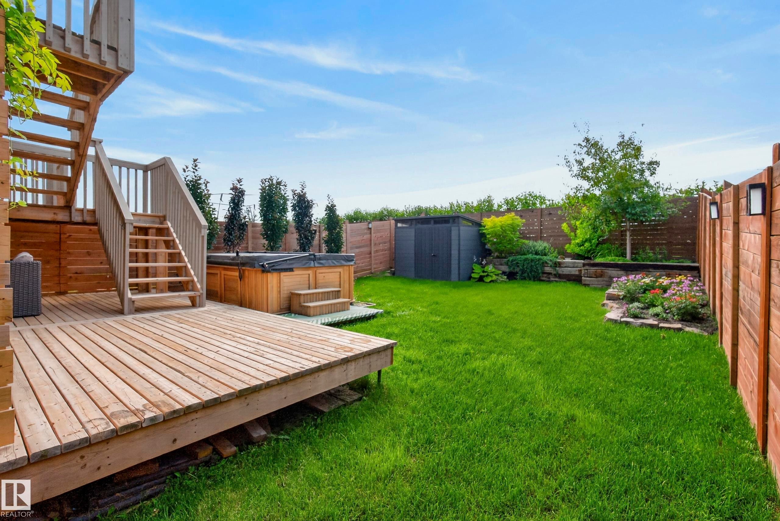 Expansive wooden deck with staircase, hot tub, lush lawn, raised garden beds, and secure privacy fencing with accent lighting - 45 Joyal Way, St. Albert, AB - Outdoor With Deck Patio Veranda