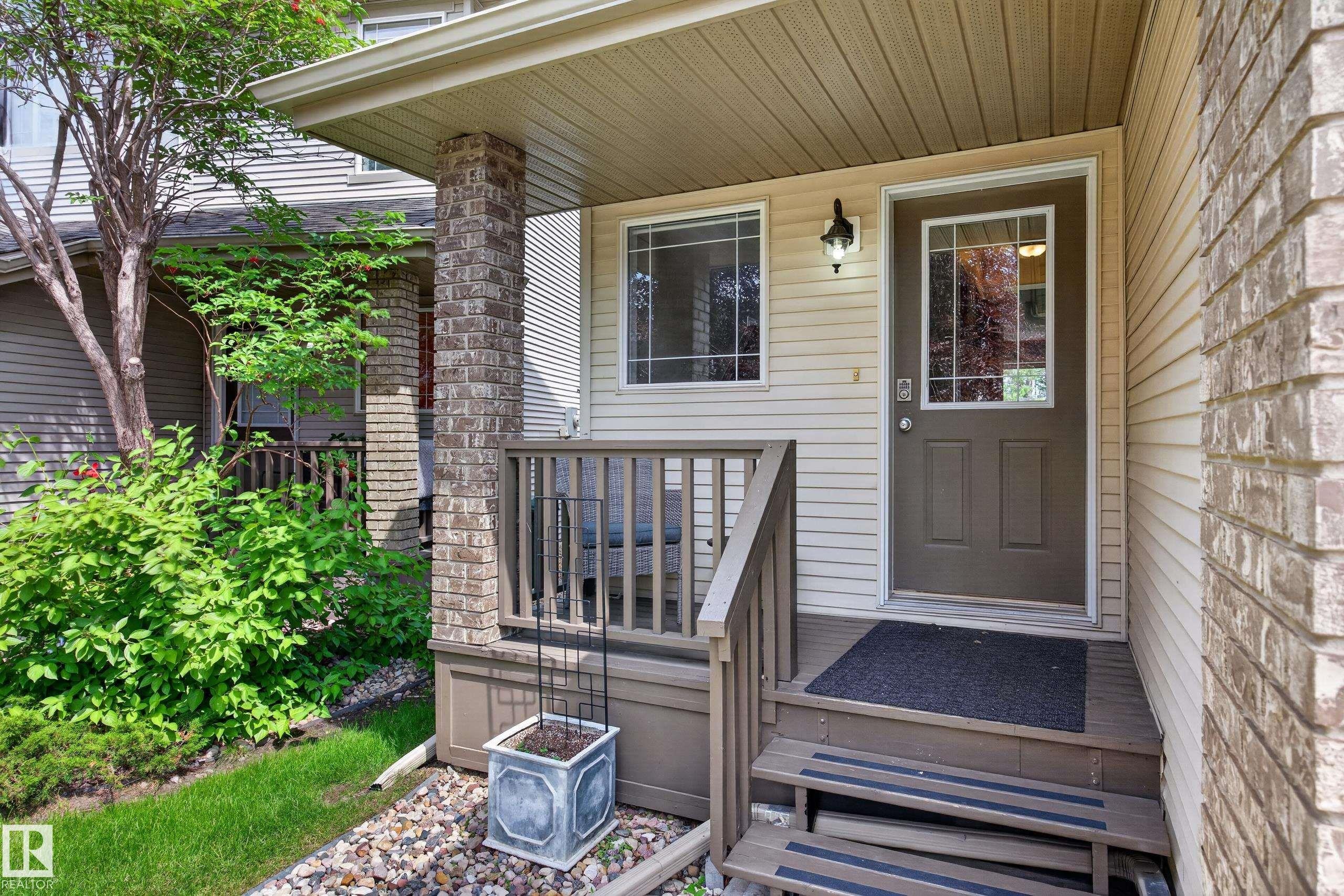 Front entrance featuring a dark brown door with decorative glass, beige siding, a brick column, a wood-toned porch with railing, and integrated exterior lighting - #7 155 Crocus Cr, Sherwood Park, AB - Outdoor With Deck Patio Veranda With Exterior