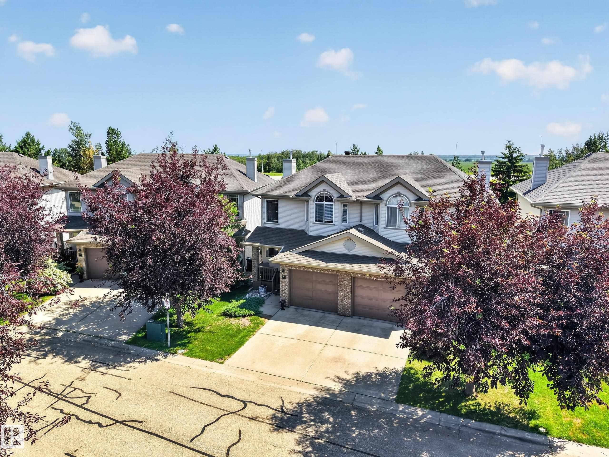 Two-story residence featuring a light-colored exterior, an attached two-car garage with wood-finish doors, a pitched roofline, and front landscaping with mature trees - #7 155 Crocus Cr, Sherwood Park, AB - Outdoor With Facade