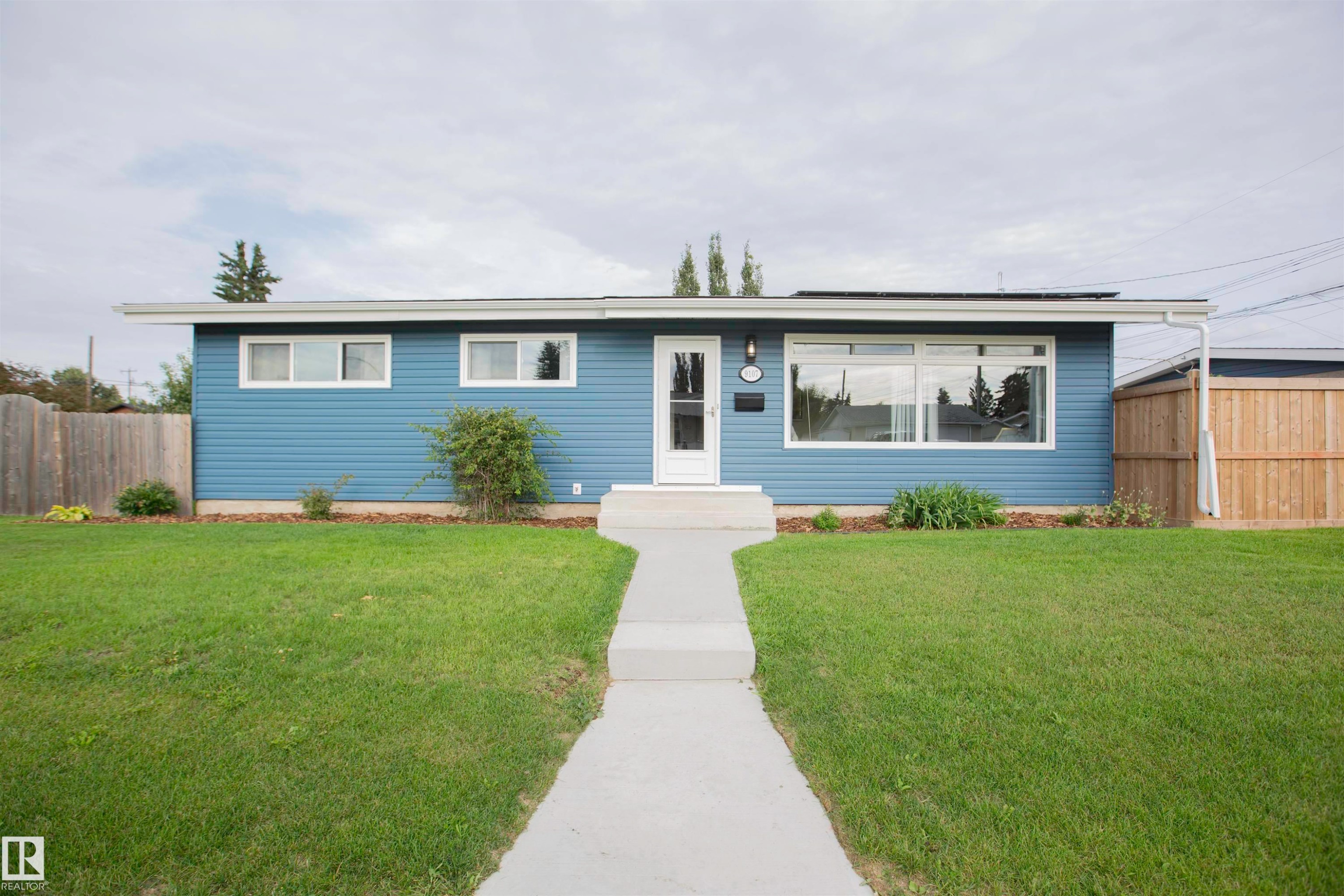 Modern ranch-style home featuring a fresh blue exterior with horizontal siding, expansive front-facing windows, and a welcoming entry with glass door - 9107 58 Street Nw, Edmonton, AB - Outdoor