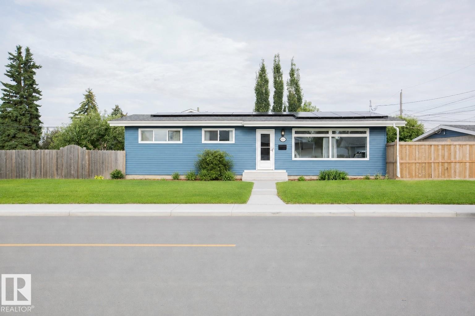 Modern blue ranch-style home featuring a clean horizontal facade with multi-pane windows, a covered carport entry, and manicured landscaping - 9107 58 Street Nw, Edmonton, AB - Outdoor With Facade