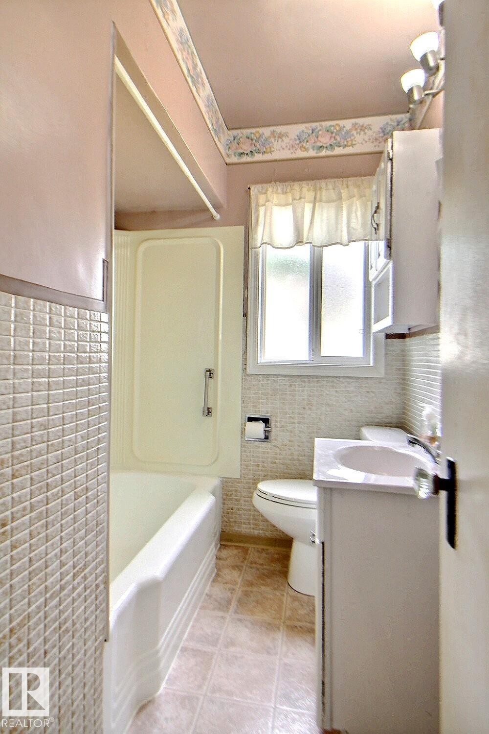 Built-in bathtub with shower enclosure, window with frosted glass, vanity with integrated sink, tiled flooring, and wall-mounted light fixture - 11336 83 Street Nw, Edmonton, AB - Indoor Photo Showing Bathroom