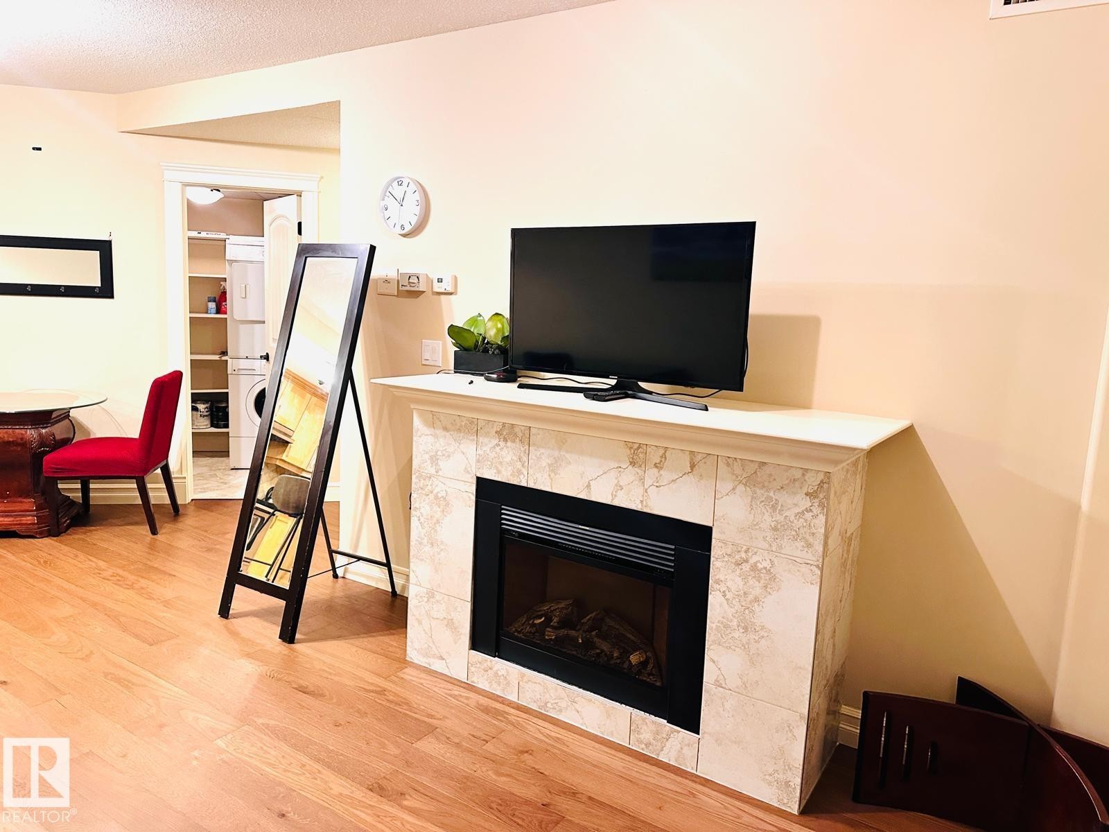 Wood-finish flooring, tiled fireplace with white mantel, and a dedicated laundry area with shelving - #207 10142 111 St Nw, Edmonton, AB - Indoor Photo Showing Living Room With Fireplace