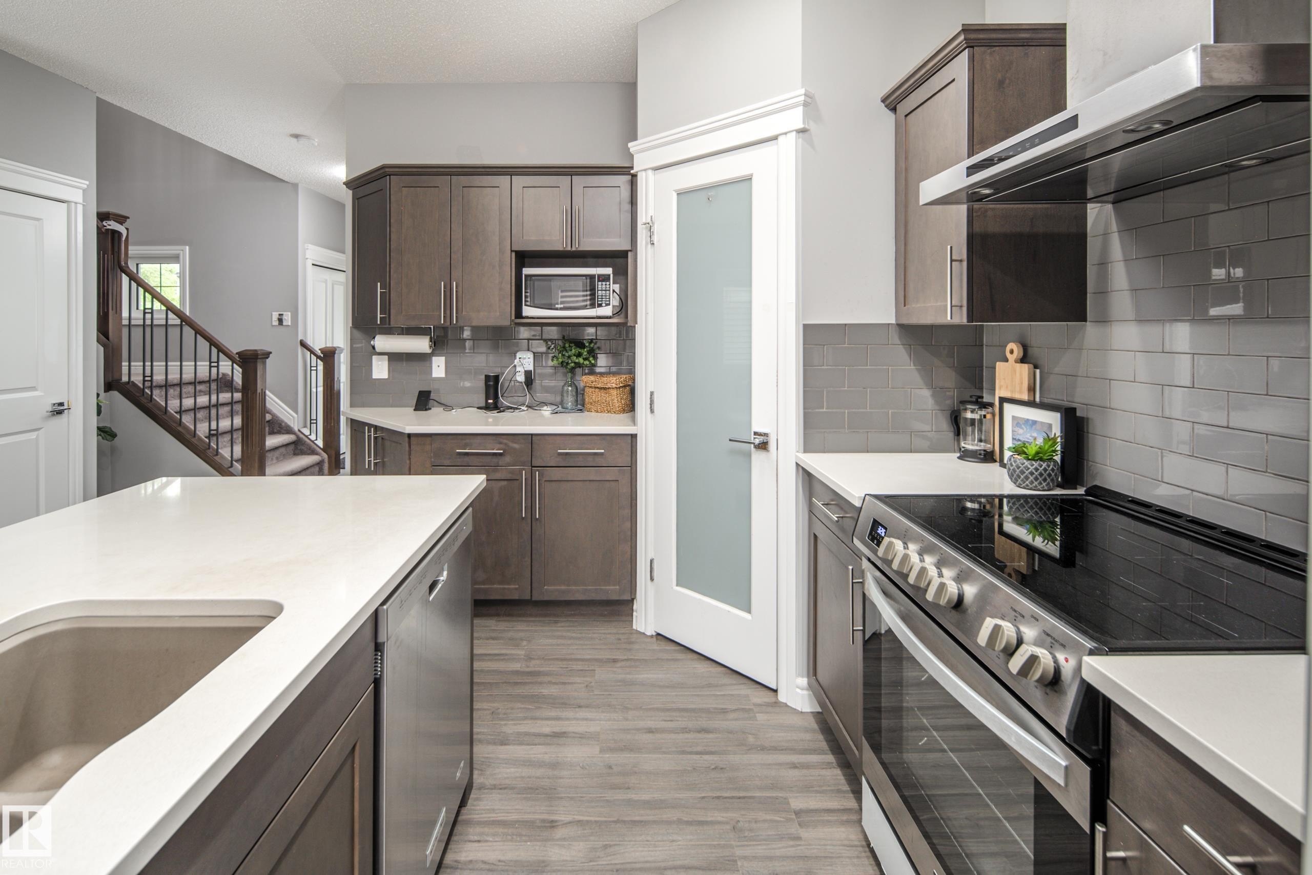 Modern kitchen featuring wood-finish flooring, stainless steel appliances, white countertops, and a gray subway tile backsplash - 16015 11 Avenue Sw, Edmonton, AB - Indoor Photo Showing Kitchen