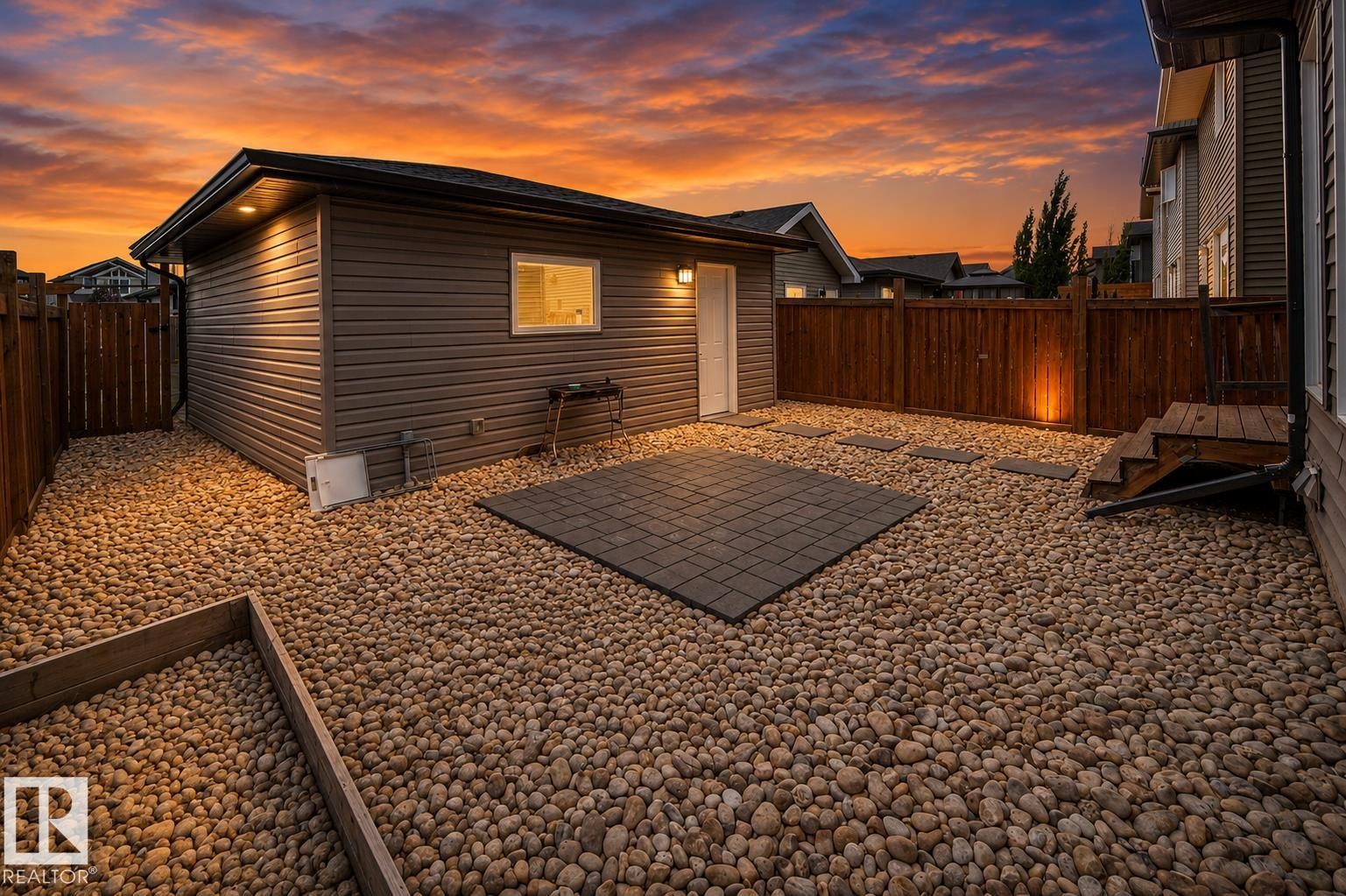 Spacious backyard retreat featuring a detached studio with illuminated interior, gravel landscaping with metal edging, composite patio pad, and wooden privacy fencing - 16015 11 Avenue Sw, Edmonton, AB - Outdoor