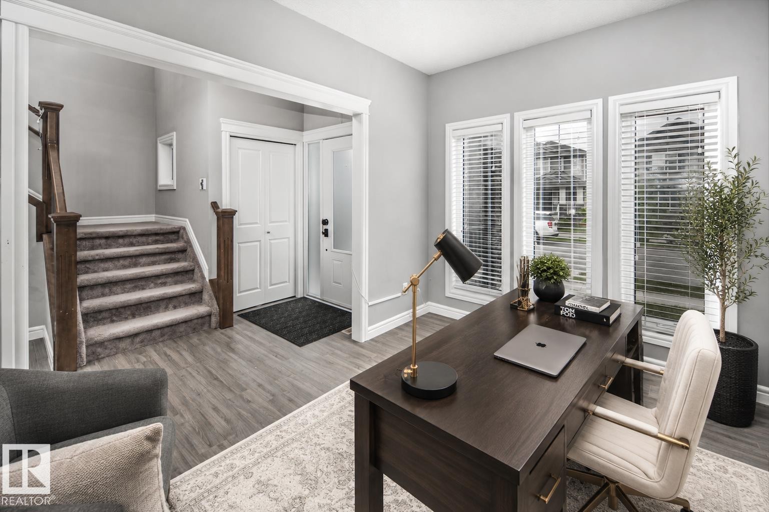 Wood-finish flooring throughout, bright entryway with a paneled door and sidelight, staircase with wood newel posts and carpeted treads, and an office space featuring three large windows with blinds - 16015 11 Avenue Sw, Edmonton, AB - Indoor Photo Showing Other Room