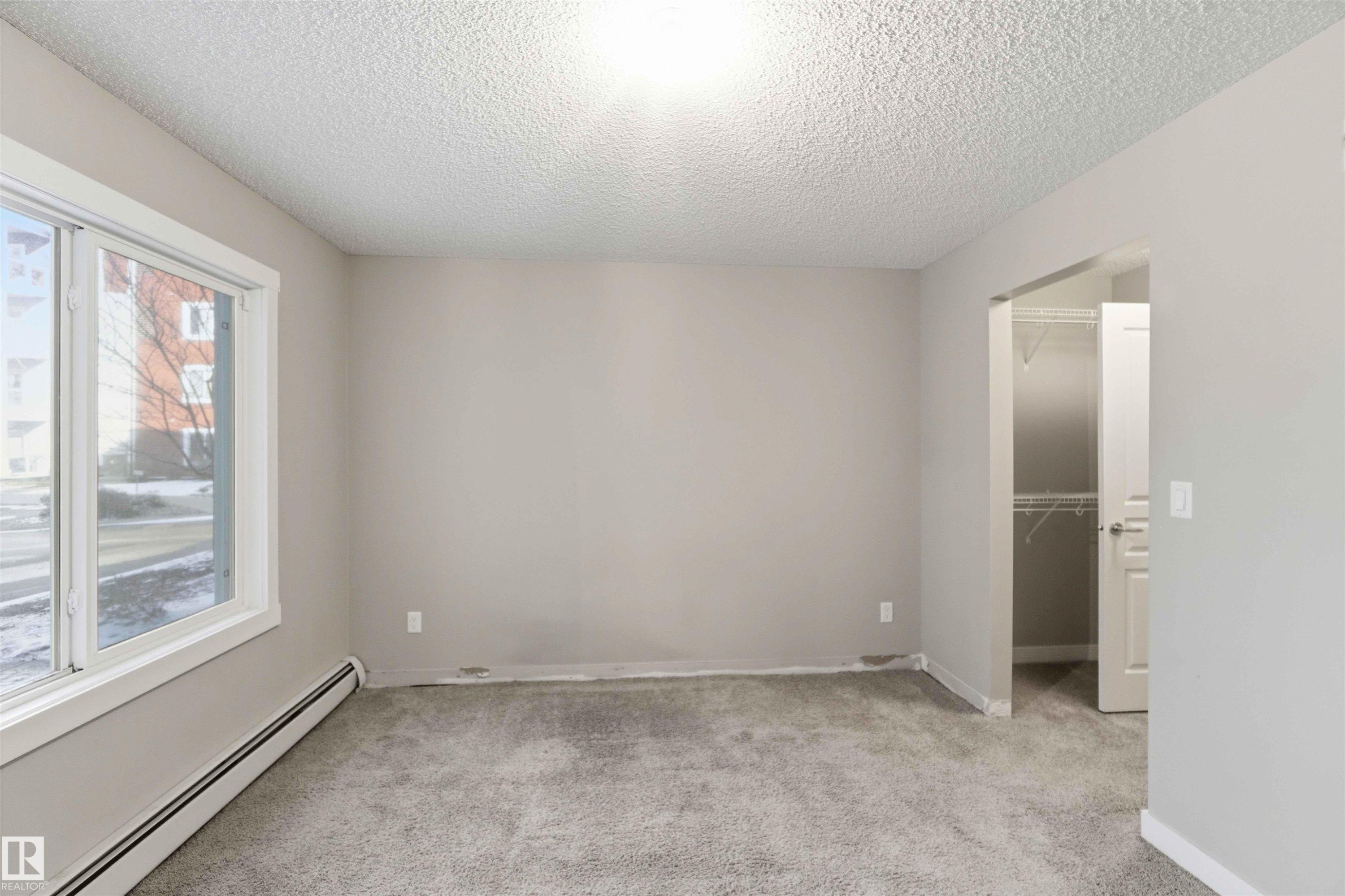 Carpeted room featuring a large window with white trim, baseboard heating, a neutral wall color, and an open doorway revealing a closet with wire shelving - #114 2207 44 Av Nw, Edmonton, AB - Indoor Photo Showing Other Room