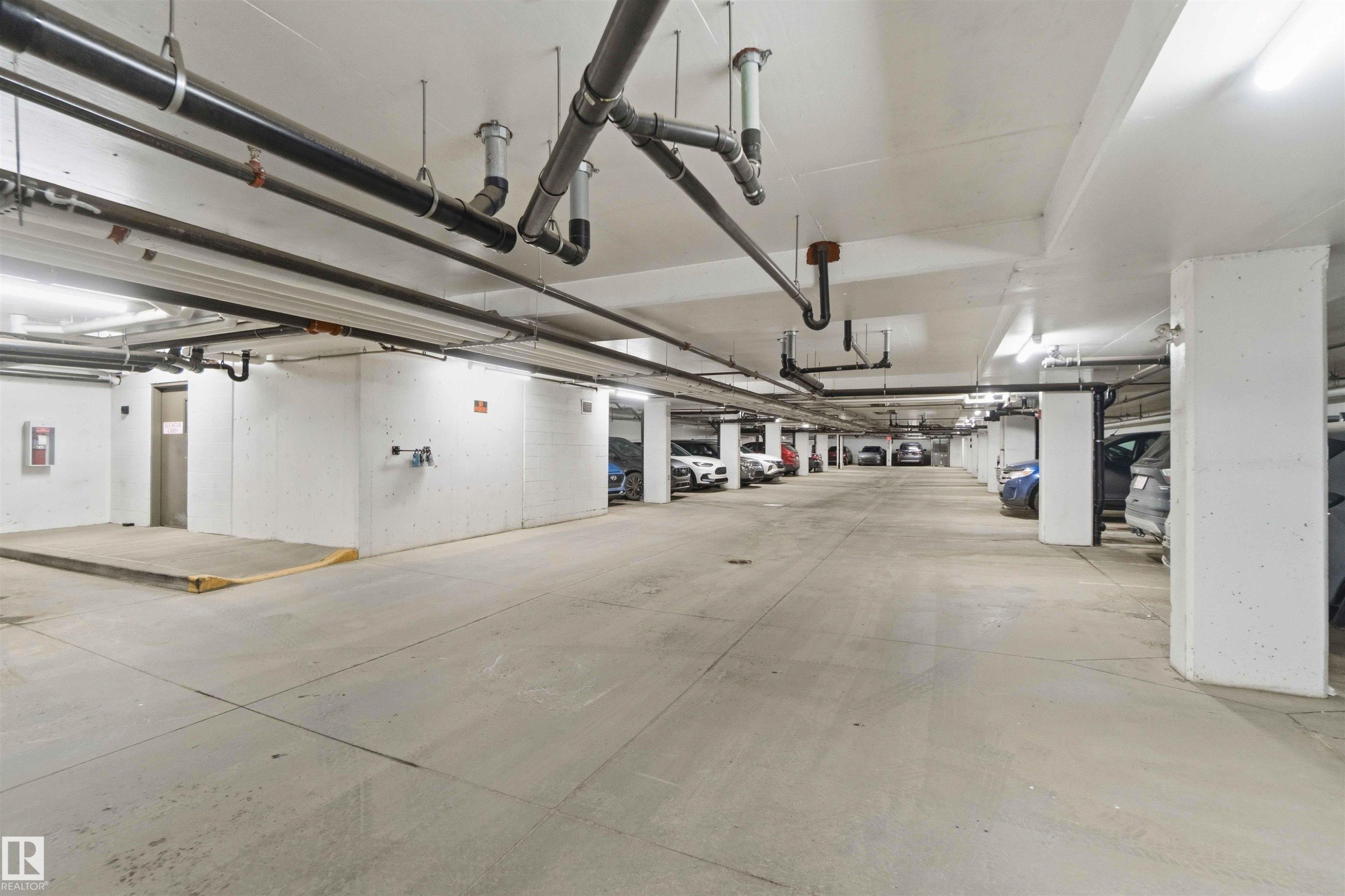 Underground parking garage featuring concrete flooring, exposed overhead piping, and fluorescent lighting - #114 2207 44 Av Nw, Edmonton, AB - Indoor Photo Showing Garage