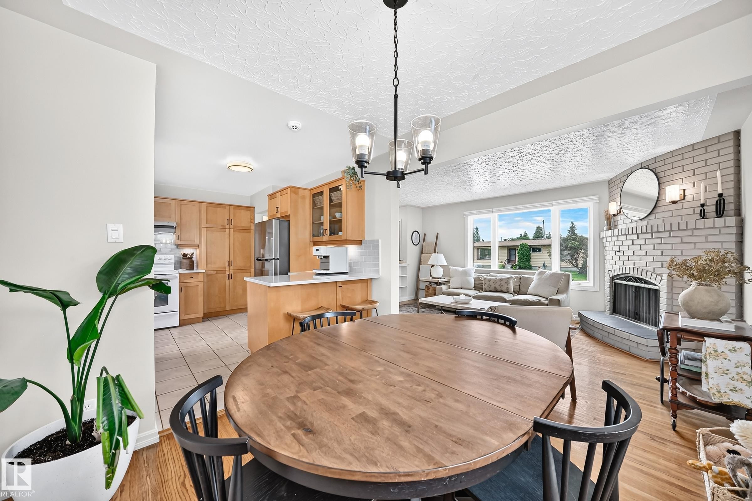 Open-concept layout with wood-finish flooring, a white brick fireplace, a large window, and an updated kitchen featuring light wood cabinetry - 16406 87A Avenue Nw, Edmonton, AB - Indoor Photo Showing Dining Room With Fireplace