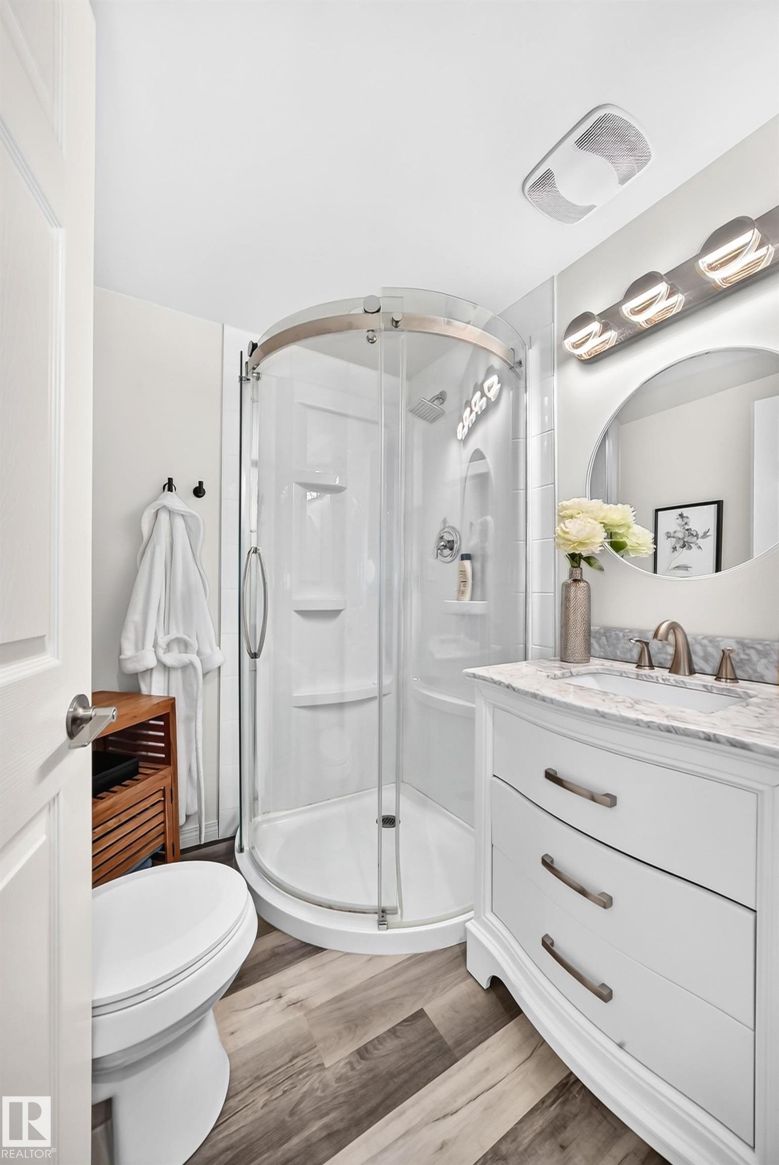 Curved glass shower enclosure, single vanity with integrated sink, stone countertop, wood-finish flooring, and wall-mounted vanity lighting - 16406 87A Avenue Nw, Edmonton, AB - Indoor Photo Showing Bathroom