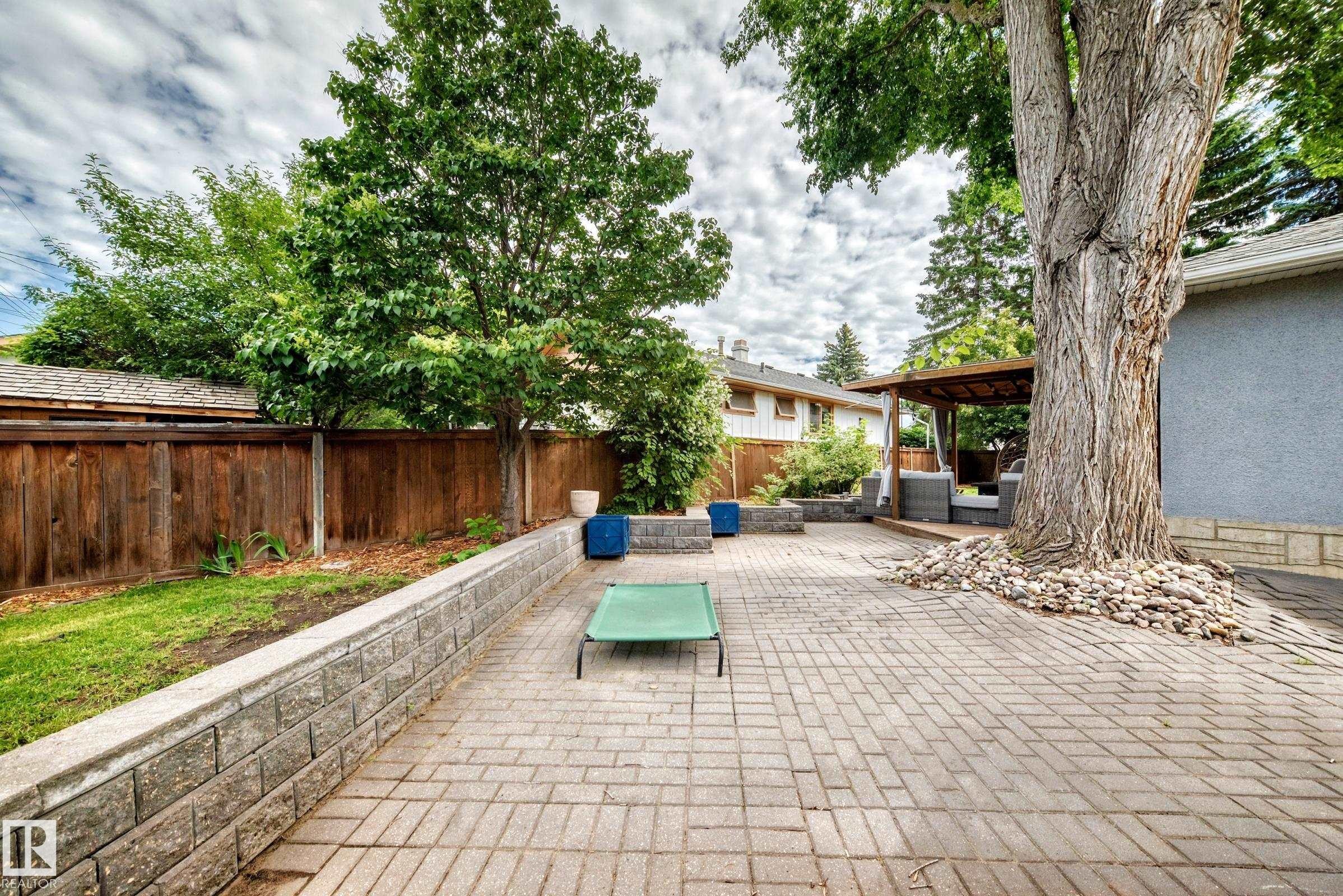 Spacious paver patio with a wood privacy fence, stone retaining wall, mature trees, and a covered outdoor seating area - 16406 87A Avenue Nw, Edmonton, AB - Outdoor