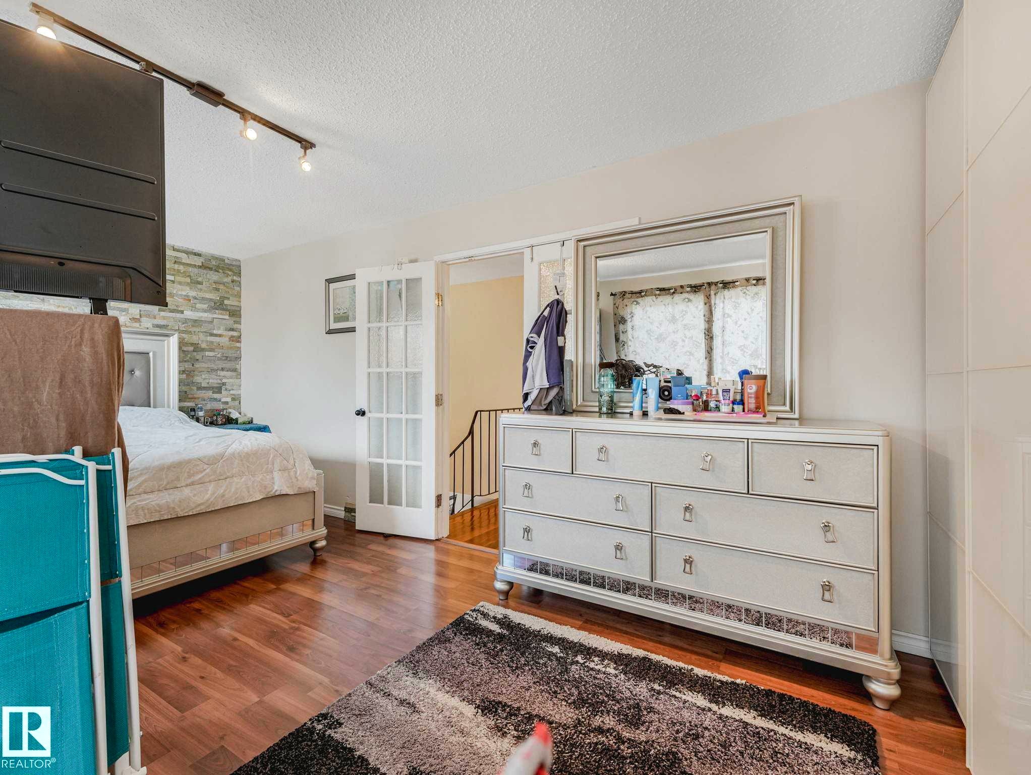 Spacious bedroom featuring wood-finish flooring, a decorative stone accent wall, track lighting, and white French doors - 13405 40 St Nw, Edmonton, AB - Indoor Photo Showing Bedroom