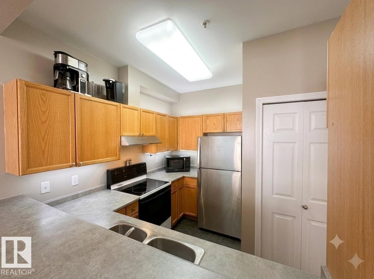 Contemporary kitchen featuring wood cabinetry, stainless steel appliances, and a double basin sink - #103 8108 109 St Nw, Edmonton, AB - Indoor Photo Showing Kitchen With Double Sink