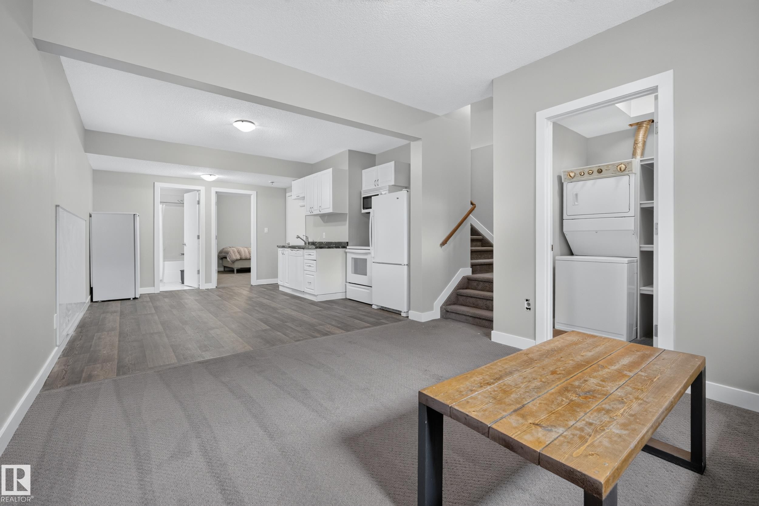 Spacious room featuring gray carpet and wood-finish flooring, a functional kitchen area with white cabinetry, a stackable washer and dryer unit, and a staircase with wood railing - 11018 122 Street Nw, Edmonton, AB - Indoor Photo Showing Other Room