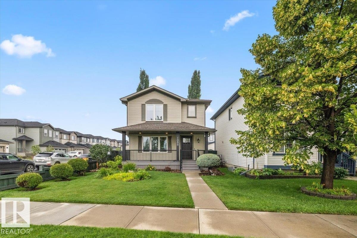 Two-story residence featuring a covered front porch with black railings, a light tan facade with dark green shutters, and a mature tree in the front yard - 1086 Grantham Drive Nw, Edmonton, AB - Outdoor With Deck Patio Veranda With Facade