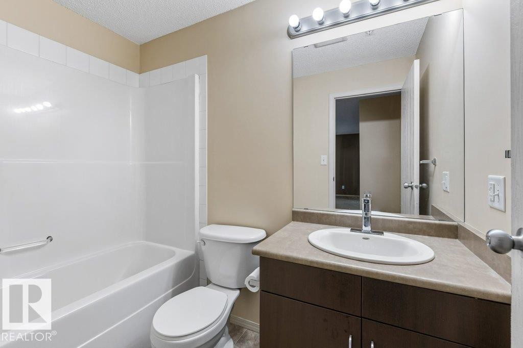 Bathroom featuring an integrated bathtub and shower, dark wood-finish vanity with a laminate countertop, and a large mirror with overhead lighting - #307 1188 Hyndman Rd Nw, Edmonton, AB - Indoor Photo Showing Bathroom