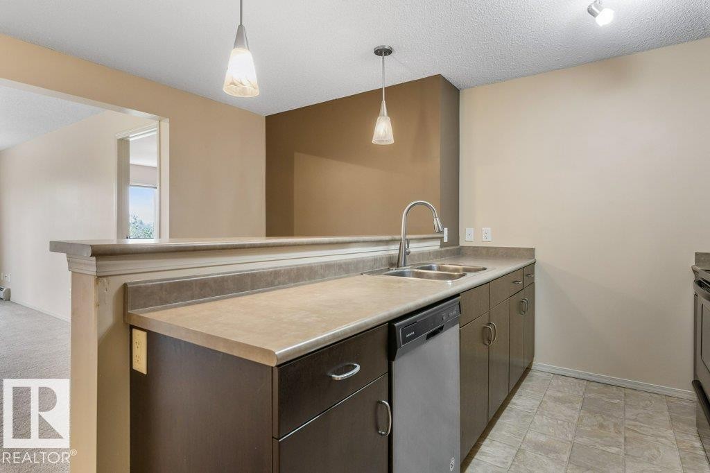 Kitchen featuring a double basin stainless steel sink, a stainless steel dishwasher, dark wood-finish cabinetry, and recessed lighting - #307 1188 Hyndman Rd Nw, Edmonton, AB - Indoor Photo Showing Kitchen With Double Sink