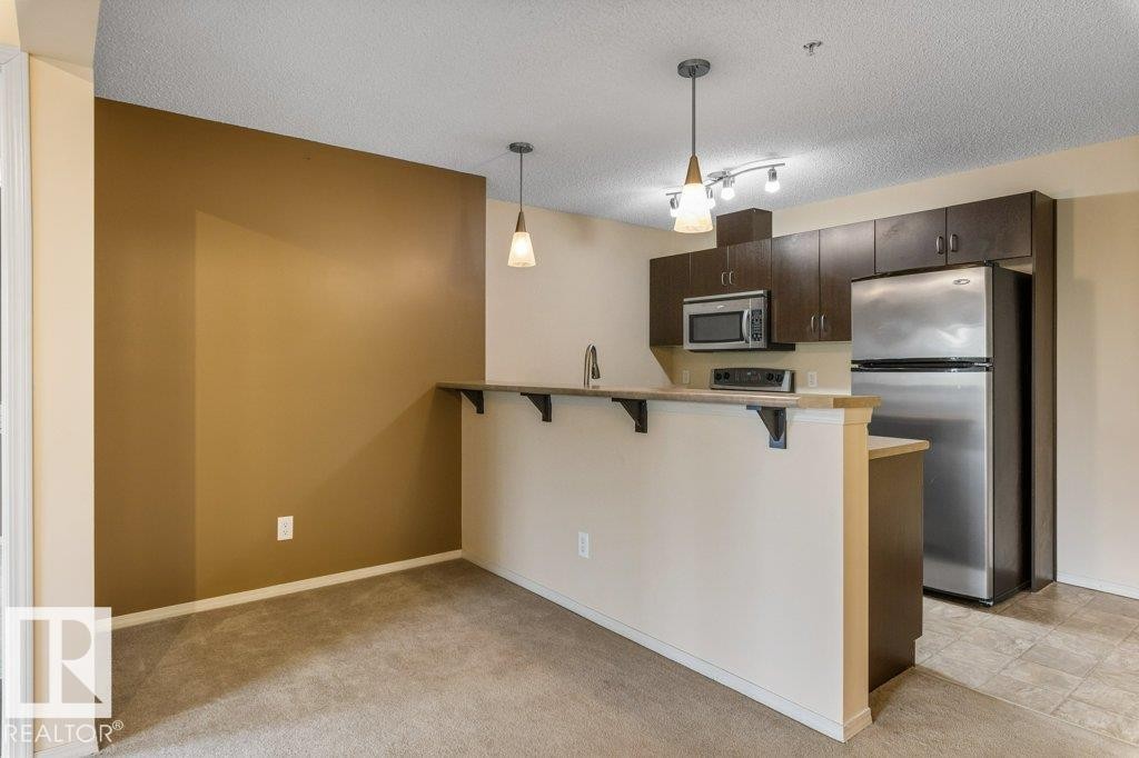 Kitchen featuring dark wood-finish cabinetry, stainless steel appliances, and a breakfast bar - #307 1188 Hyndman Rd Nw, Edmonton, AB - Indoor Photo Showing Kitchen