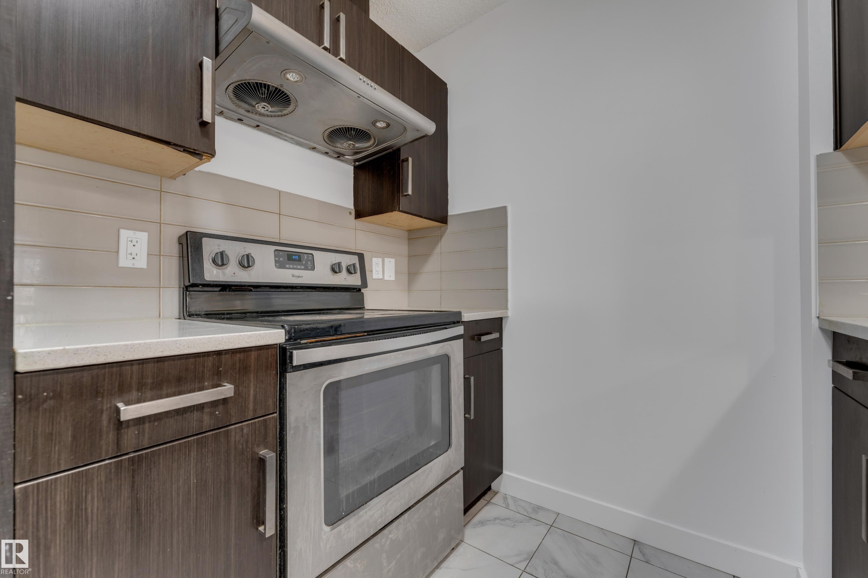 Dark wood-finish cabinetry, stainless steel range with overhead ventilation, light-toned countertop, horizontal tile backsplash, and tile flooring - #217 667 Watt Bv Sw, Edmonton, AB - Indoor Photo Showing Kitchen