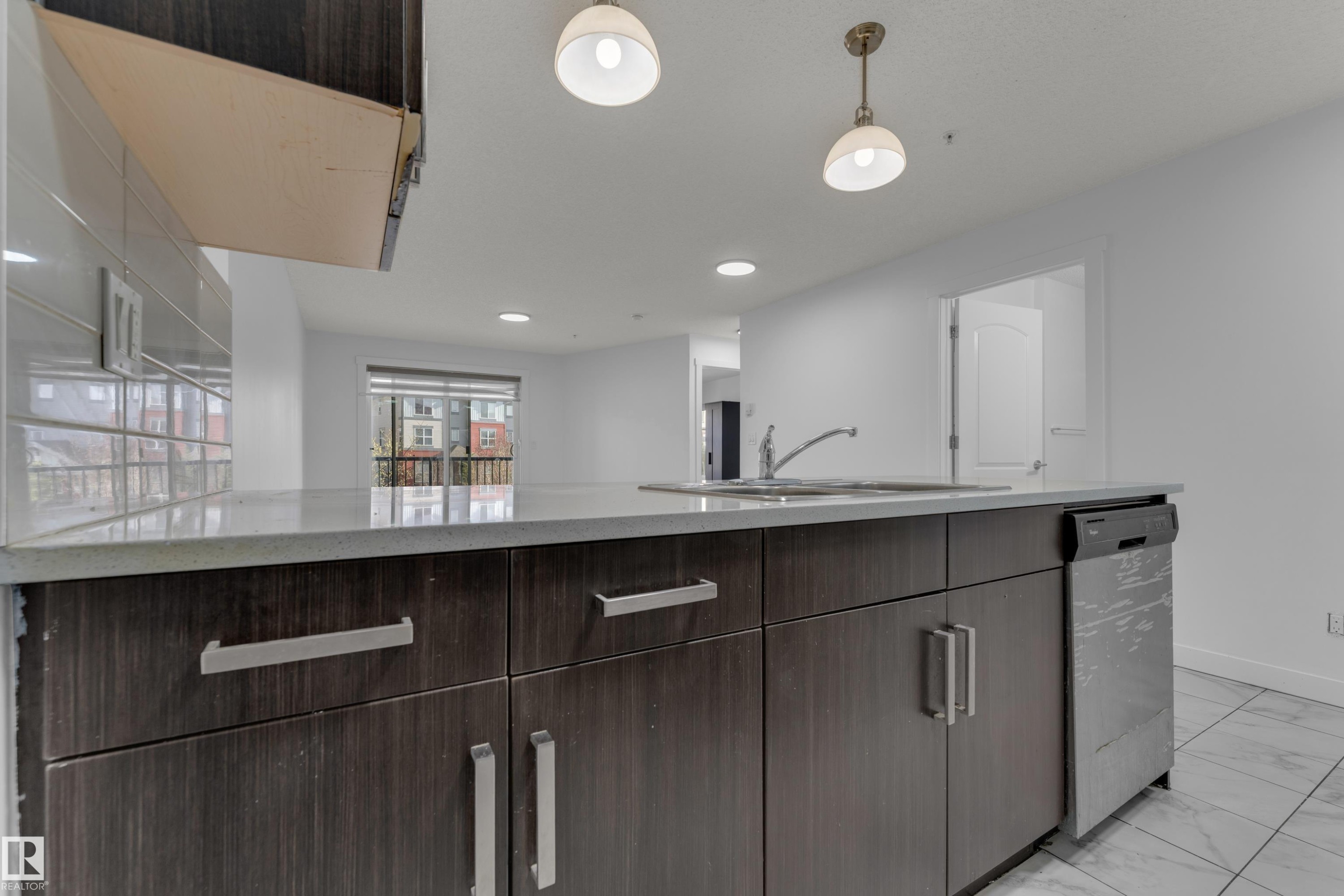 Kitchen island featuring dark wood-finish cabinetry, light-toned countertops, an undermount sink with a gooseneck faucet, a built-in stainless steel dishwasher, and two pendant light fixtures - #217 667 Watt Bv Sw, Edmonton, AB - Indoor Photo Showing Kitchen