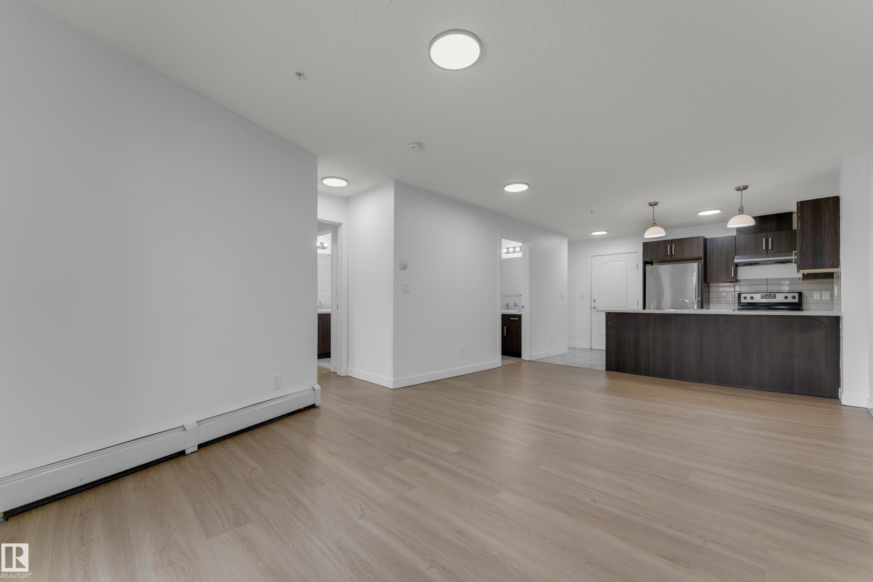 Open-concept living area with wood-finish flooring, recessed lighting, and a kitchen featuring dark wood cabinetry, stainless steel appliances, and a breakfast bar with pendant lighting - #217 667 Watt Bv Sw, Edmonton, AB - Indoor Photo Showing Kitchen