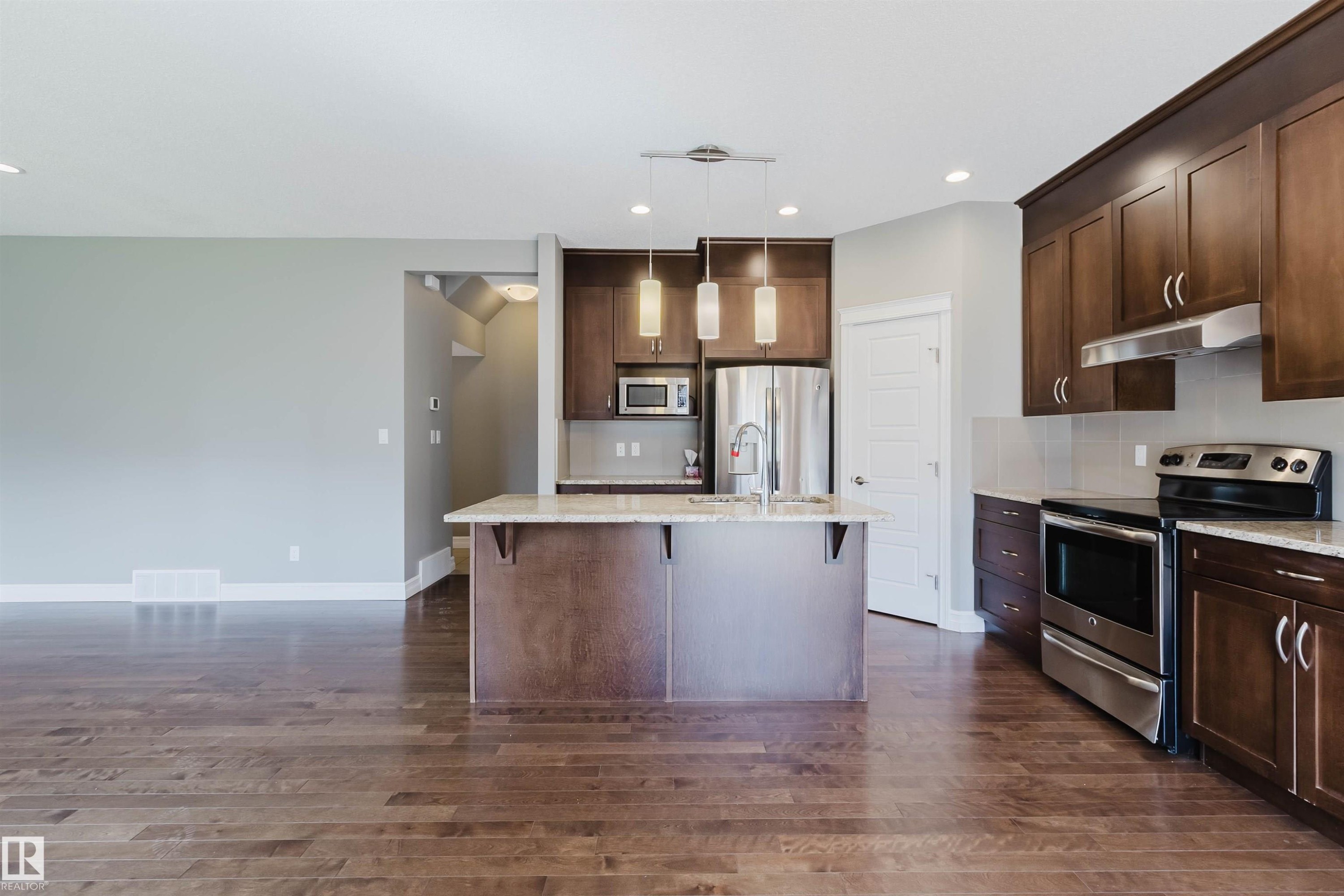 Kitchen featuring wood-finish flooring, dark wood cabinetry, stainless steel appliances, a center island with pendant lighting, and light-colored countertops - 15839 10 Avenue Sw, Edmonton, AB - Indoor Photo Showing Kitchen With Stainless Steel Kitchen With Upgraded Kitchen