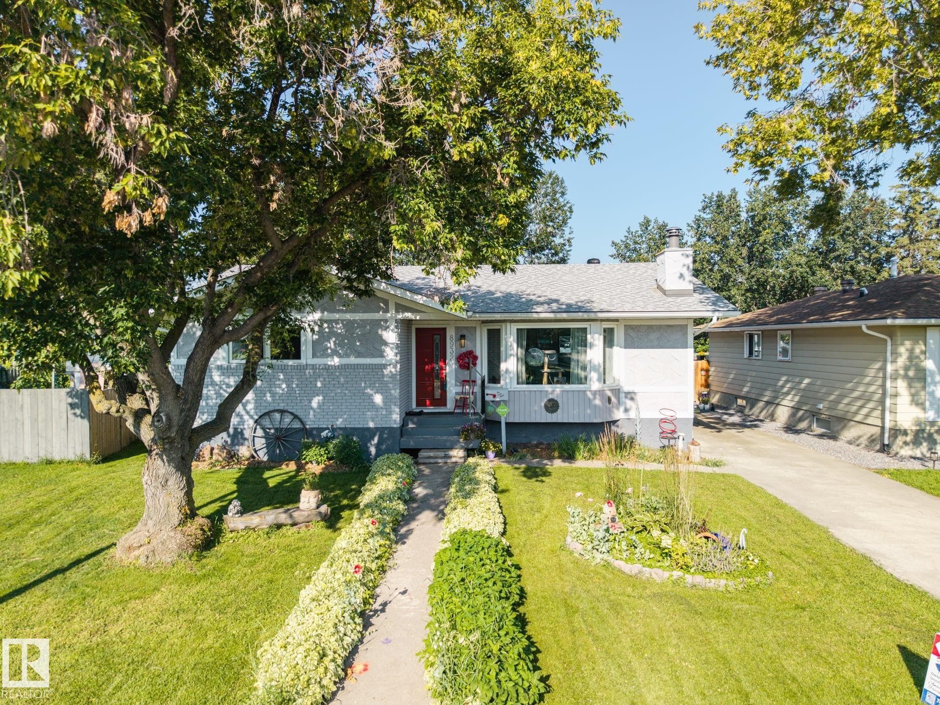 Exterior featuring a light-colored facade, a prominent bay window, a red entry door, a paved driveway, and a front yard with mature landscaping - 8932 187 Street Nw, Edmonton, AB - Outdoor