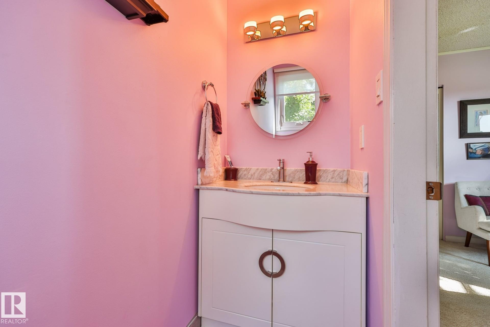 Stylish powder room featuring a soft pink accent wall with integrated LED lighting, round mirror with chrome frame, white cabinetry with circular hardware detail, and a natural wood-finish countertop with chrome faucet and sink fixtures - 8932 187 Street Nw, Edmonton, AB - Indoor Photo Showing Bathroom