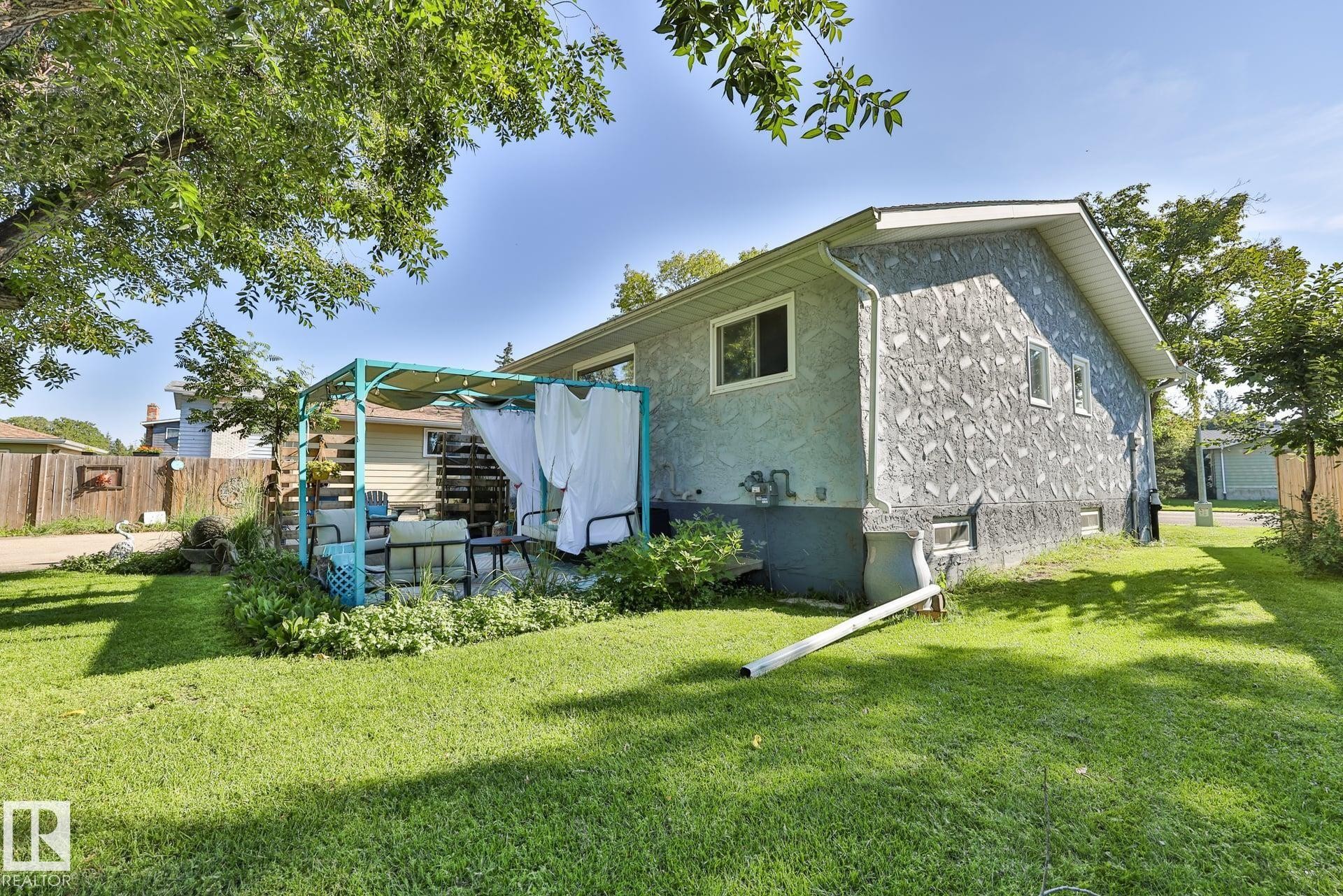 Expansive lawn area with mature trees, a stucco exterior, and a dedicated patio space featuring a pergola - 8932 187 Street Nw, Edmonton, AB - Outdoor