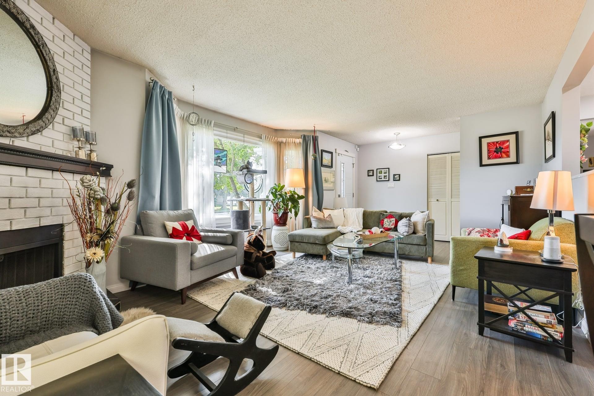 Spacious living area featuring a white brick fireplace with a dark wood mantel, wood-finish flooring, and ample natural light from a large window - 8932 187 Street Nw, Edmonton, AB - Indoor Photo Showing Living Room With Fireplace
