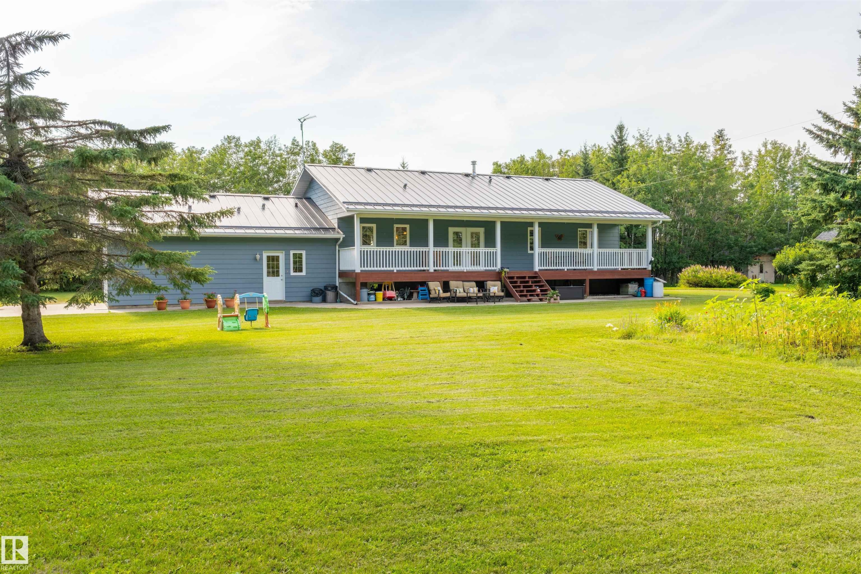 Expansive lawn with mature trees, featuring a light blue siding residence with a metal roof and a full-width covered porch - 59125 Range Road 102, Rural St. Paul County, AB - Outdoor With Deck Patio Veranda