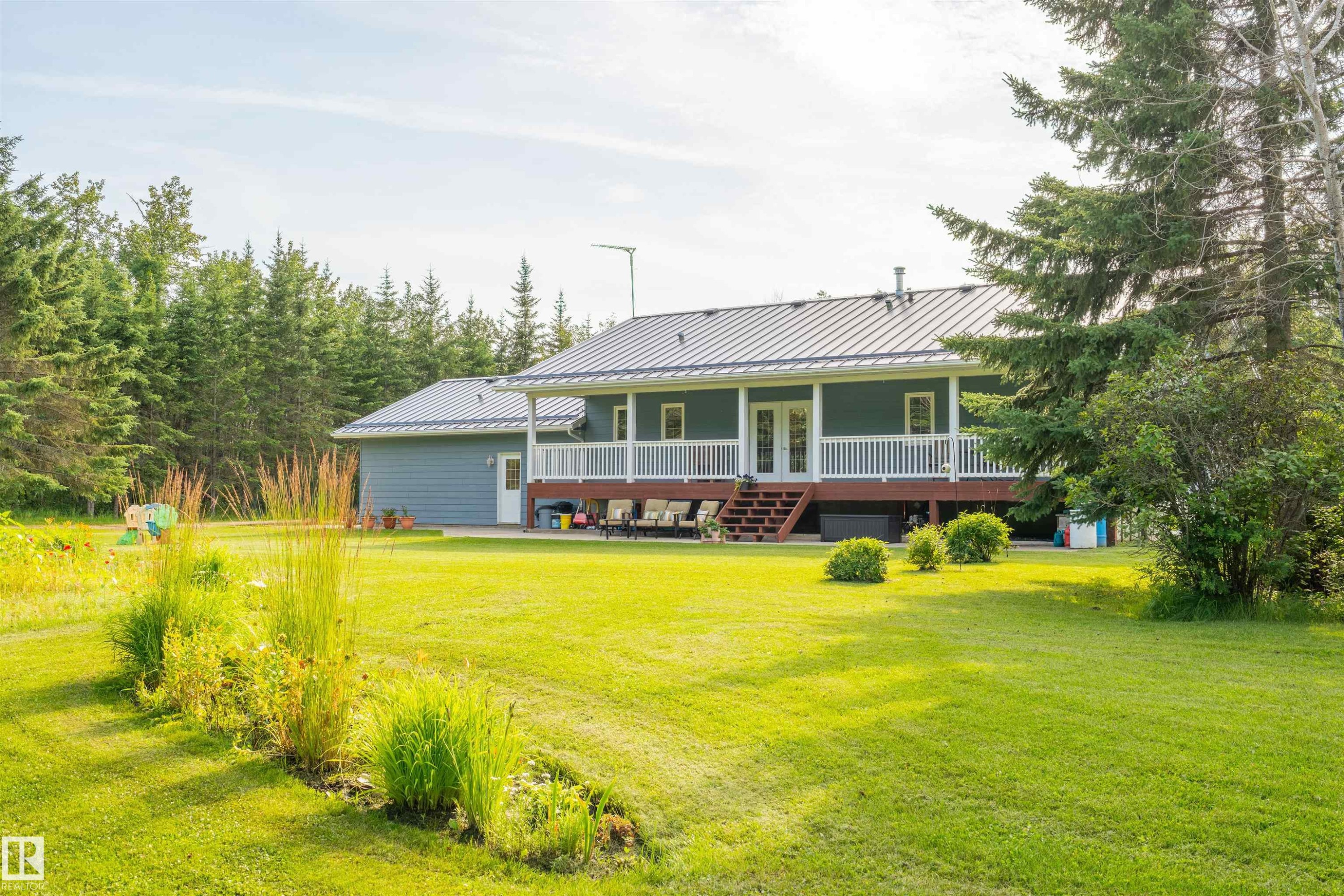 Expansive green lawn surrounds a property featuring a metal roof, a full-length covered deck with white railings, and an attached garage - 59125 Range Road 102, Rural St. Paul County, AB - Outdoor With Deck Patio Veranda